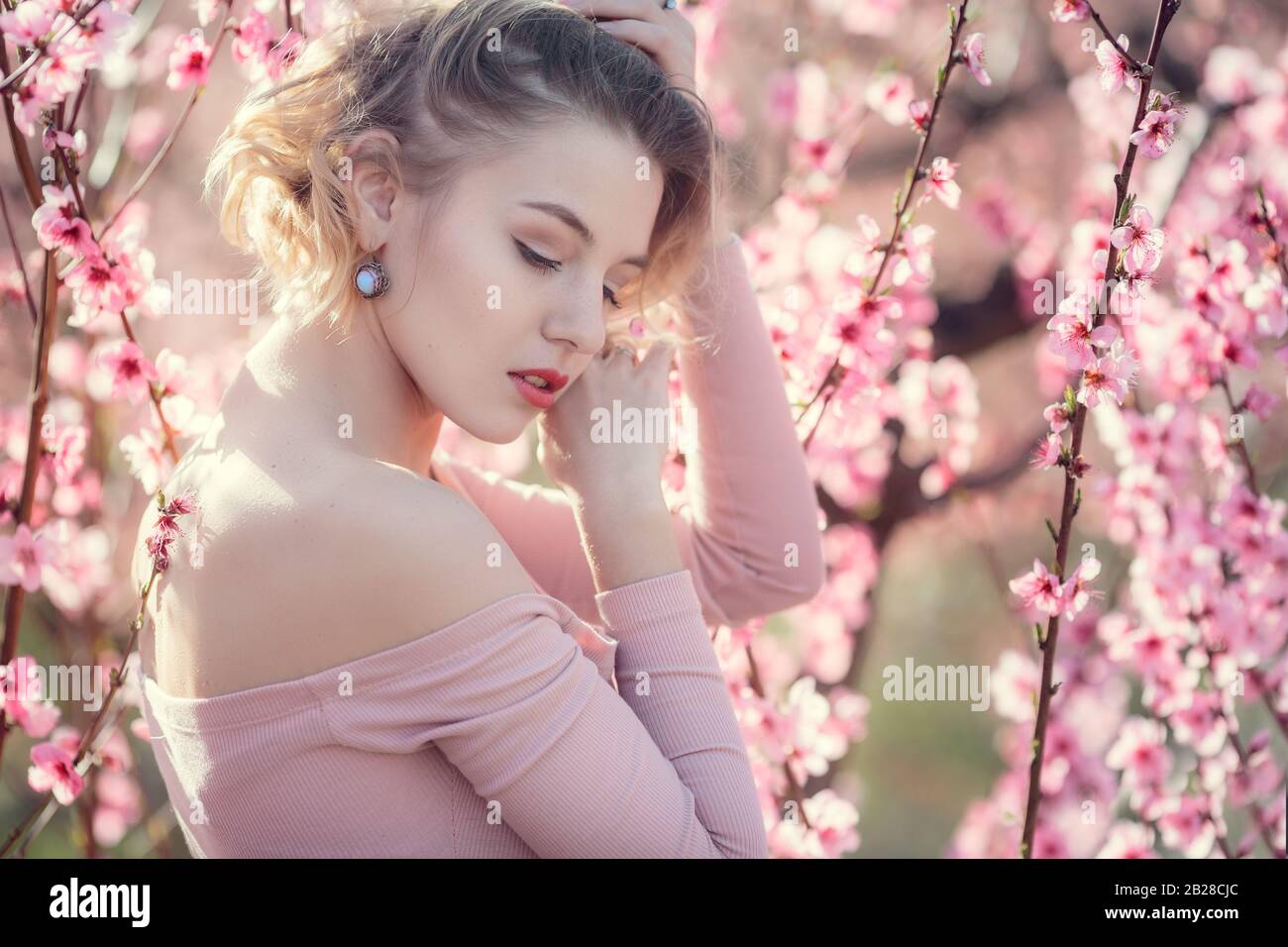 fashion outdoor photo of gorgeous young woman in elegant dress posing in garden with blossom peach trees. Blonde in flowering gardens Stock Photo