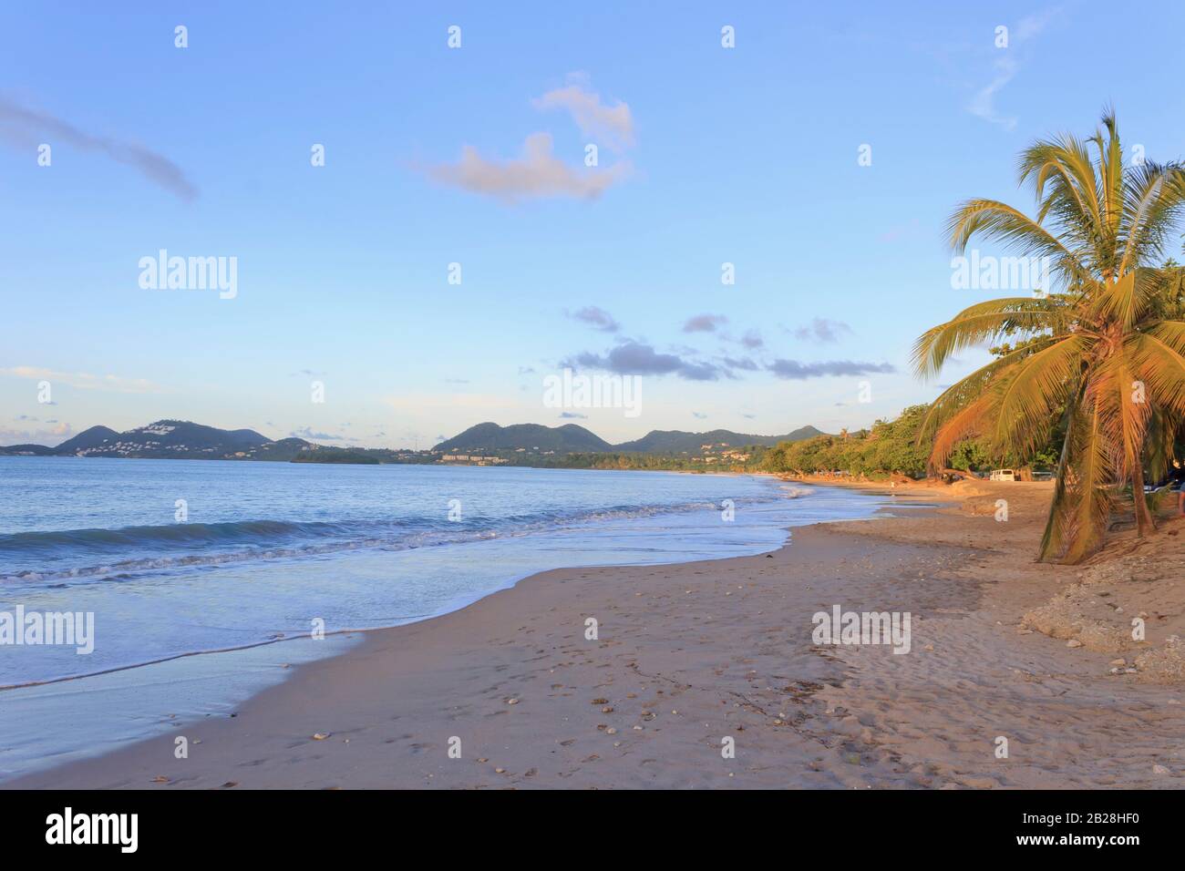 Vigie beach at sunrise, gentle wave from the blue sea, the green covered hills yonder, palm and almond trees lit yellow from the rising sun Stock Photo