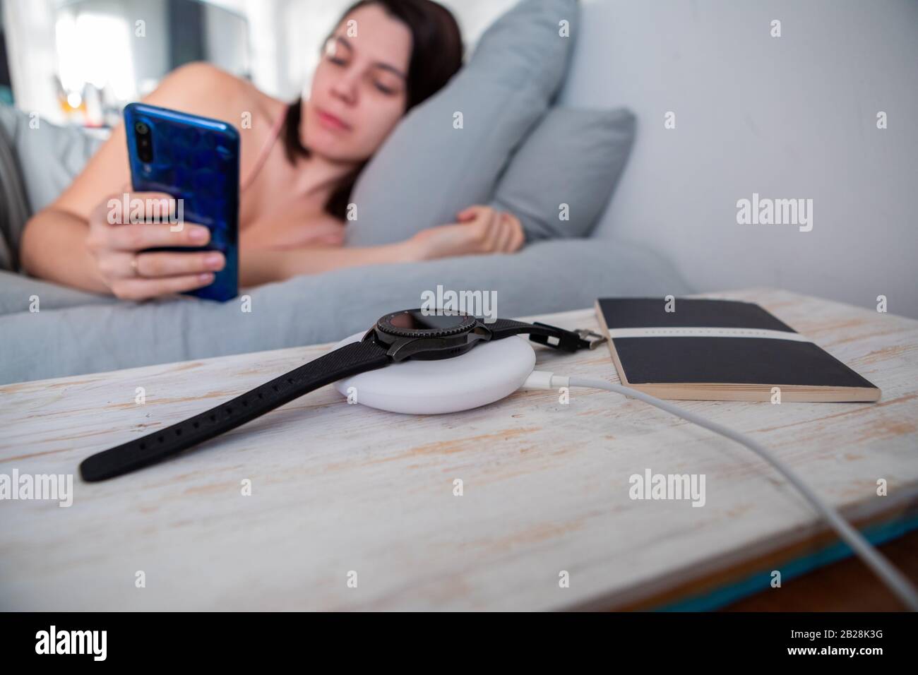 woman surfing internet on cellphone laying in bed smart watch on wireless charger Stock Photo