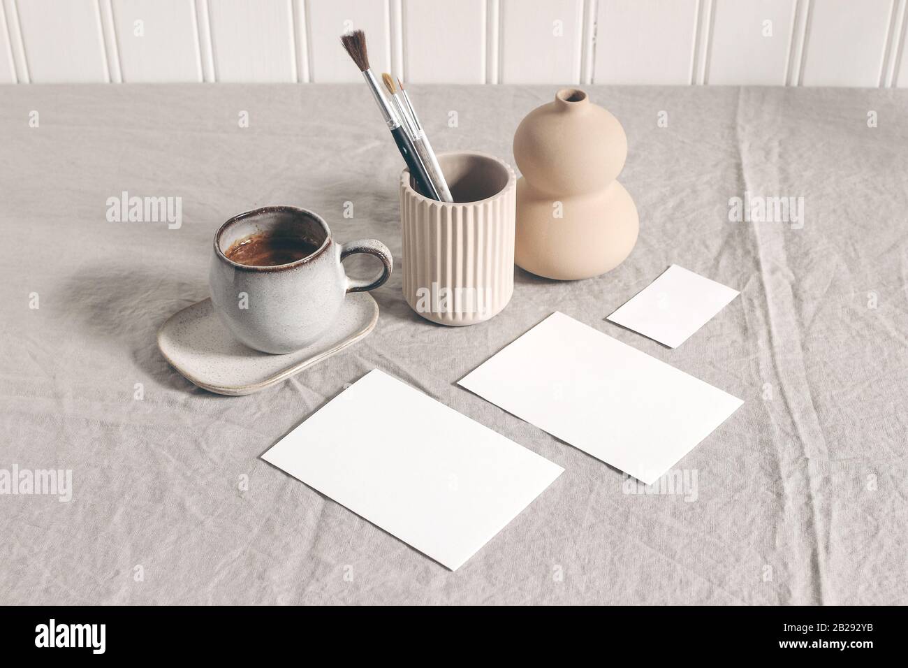 Artistic wokspace, still life. Paint brushes, pencils in ceramic holder, vase, cup of coffee and blank paper card mockups on linen tablecloth. Art Stock Photo