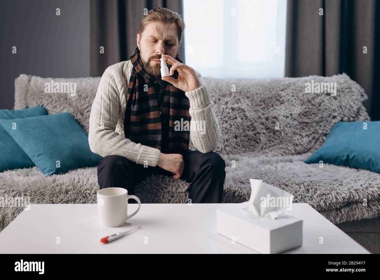 Mature sick man in warm home clothing and knitted scarf sitting on sofa at  home near table with medicines. Bearded male using nasal spray to make brea  Stock Photo - Alamy