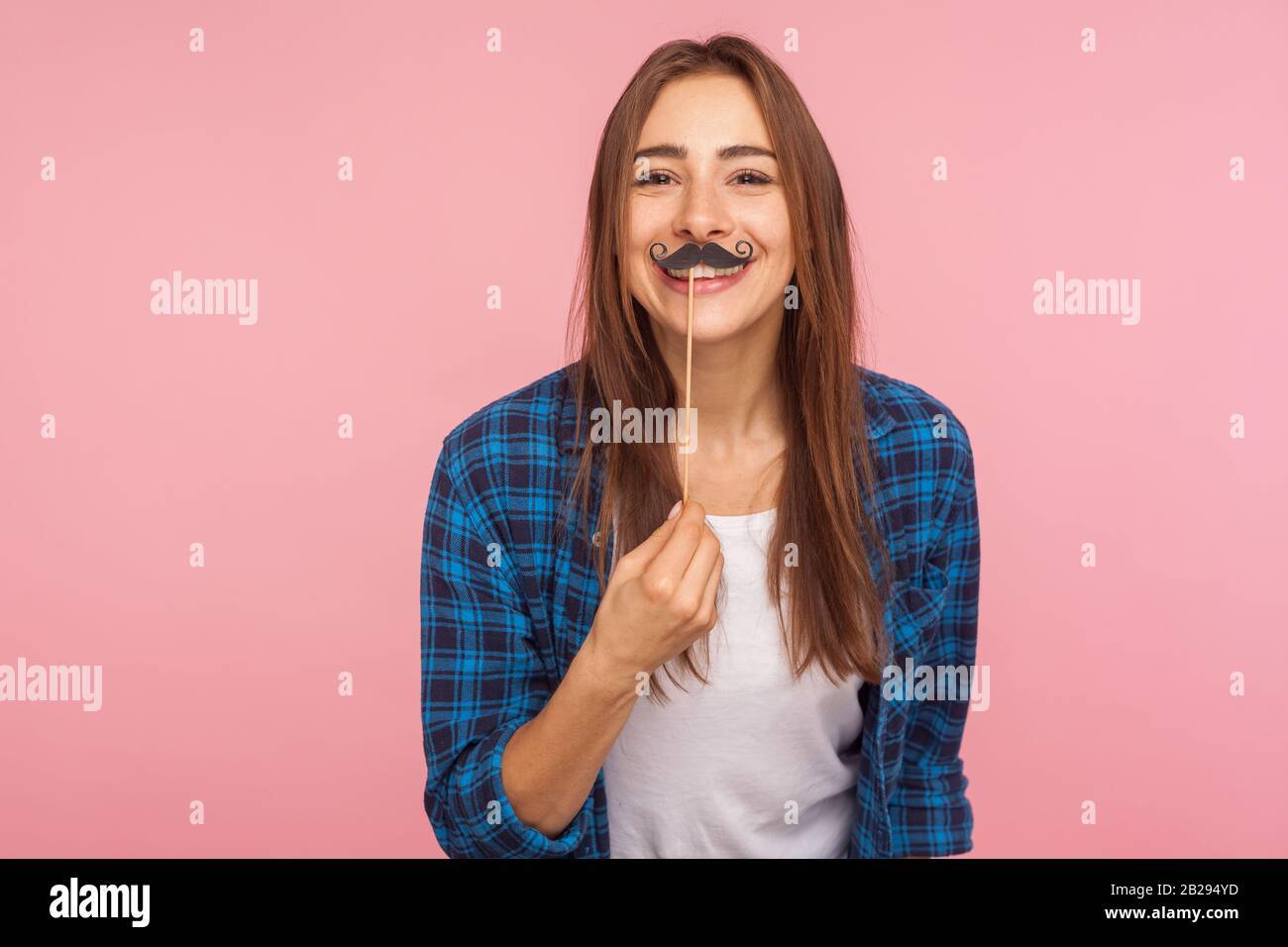Portrait of cheerful playful girl in checkered shirt holding fake curled mustache on stick and smiling to camera, having fun, wearing masquerade acces Stock Photo