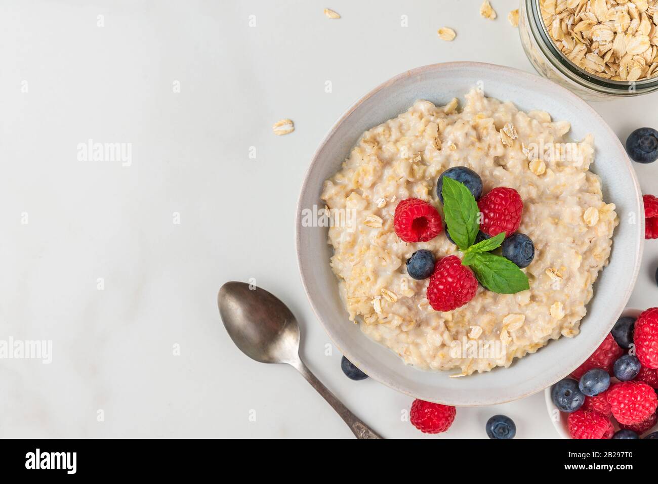 Bowl of oatmeal porridge with fresh berries and mint served with a spoon for healthy diet breakfast on white table. top view with copy space Stock Photo