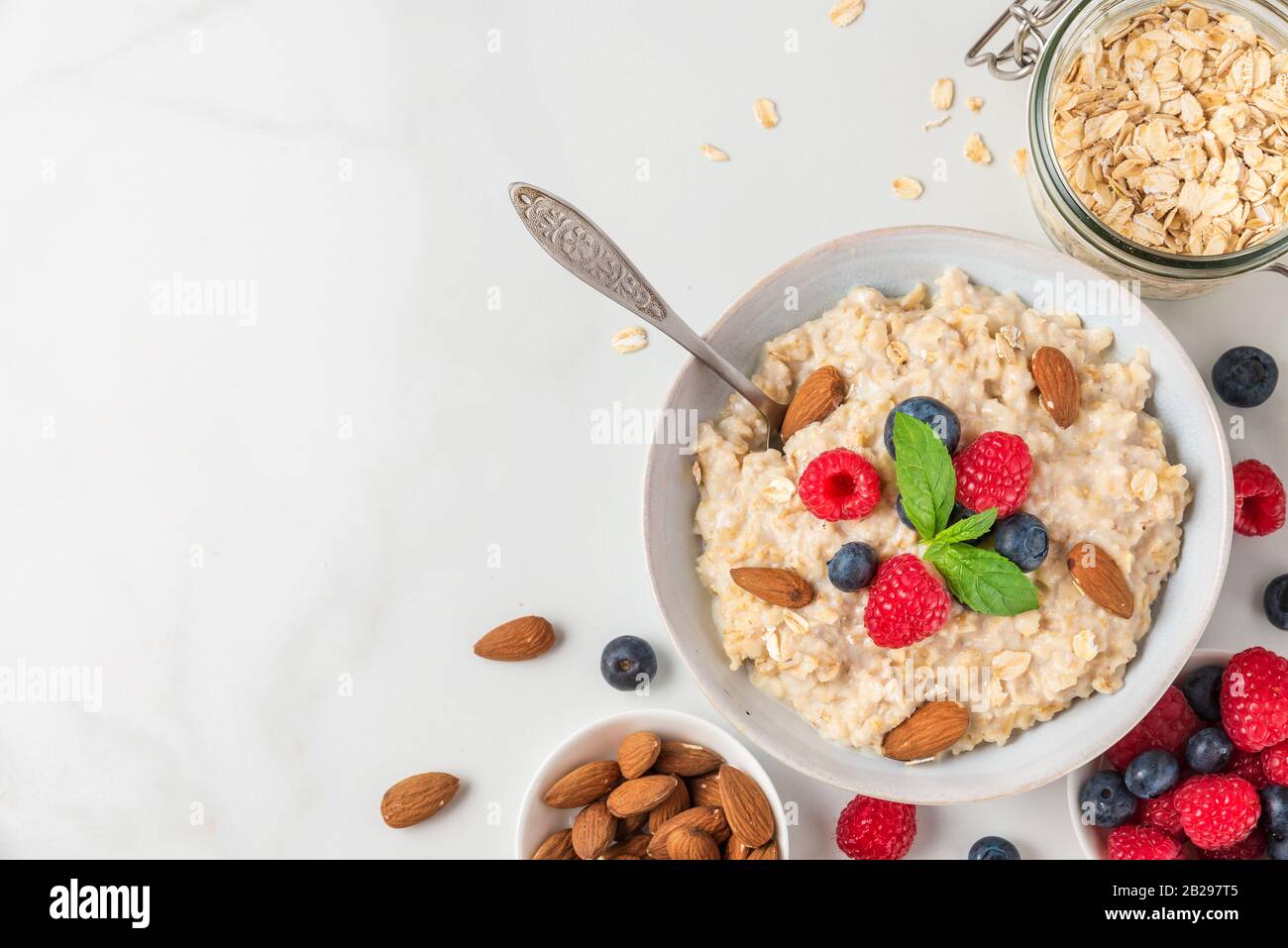 Bowl of oatmeal porridge with fresh berries, almonds and mint with a spoon for healthy diet breakfast on white table. top view with copy space Stock Photo