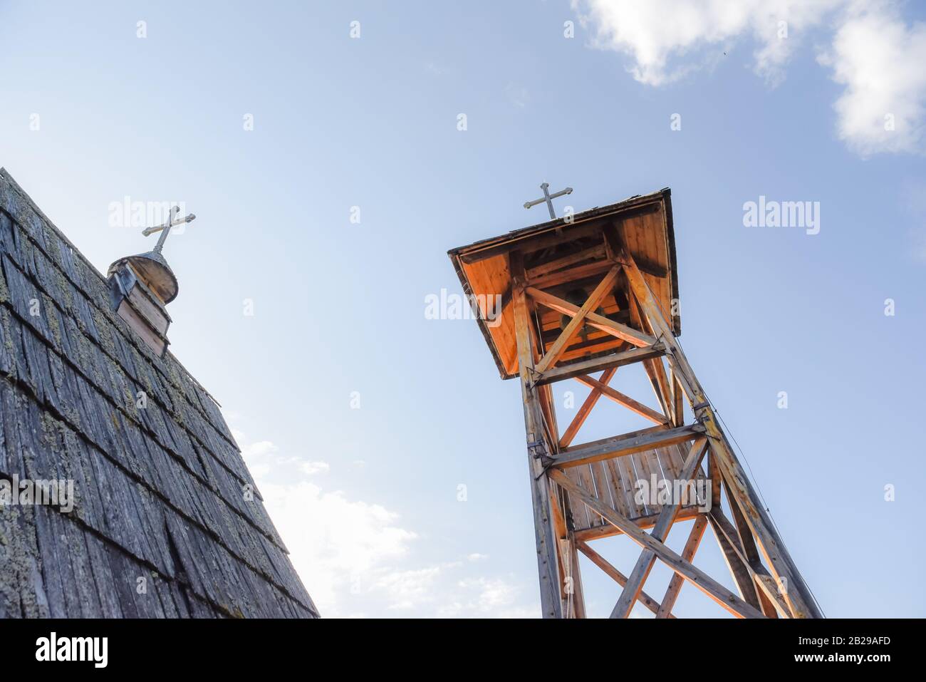 Wooden hut roof built from hay with a cross on top as medieval church in Serbia on a sunny day with a wooden bell tower with brass bell beside it Stock Photo