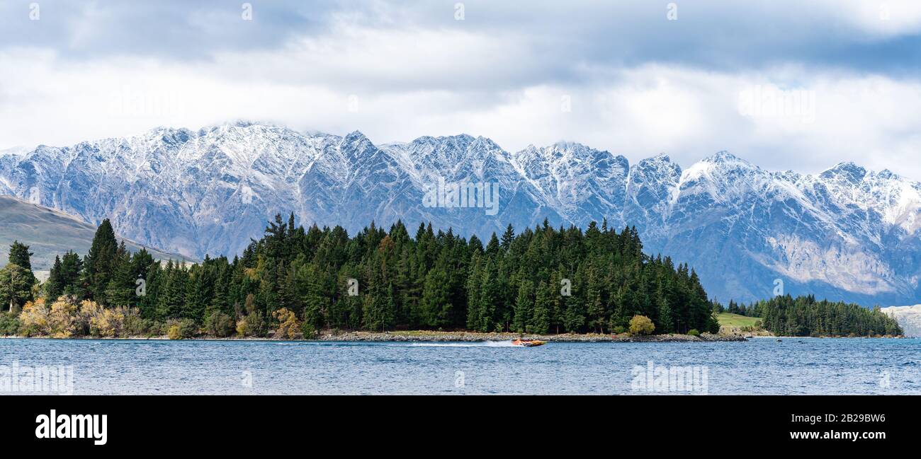 A jet boat full of joy riders seeking a thrill along the shores of Lake Wakatipu wiith a forest and snowy alpine mountain background Stock Photo