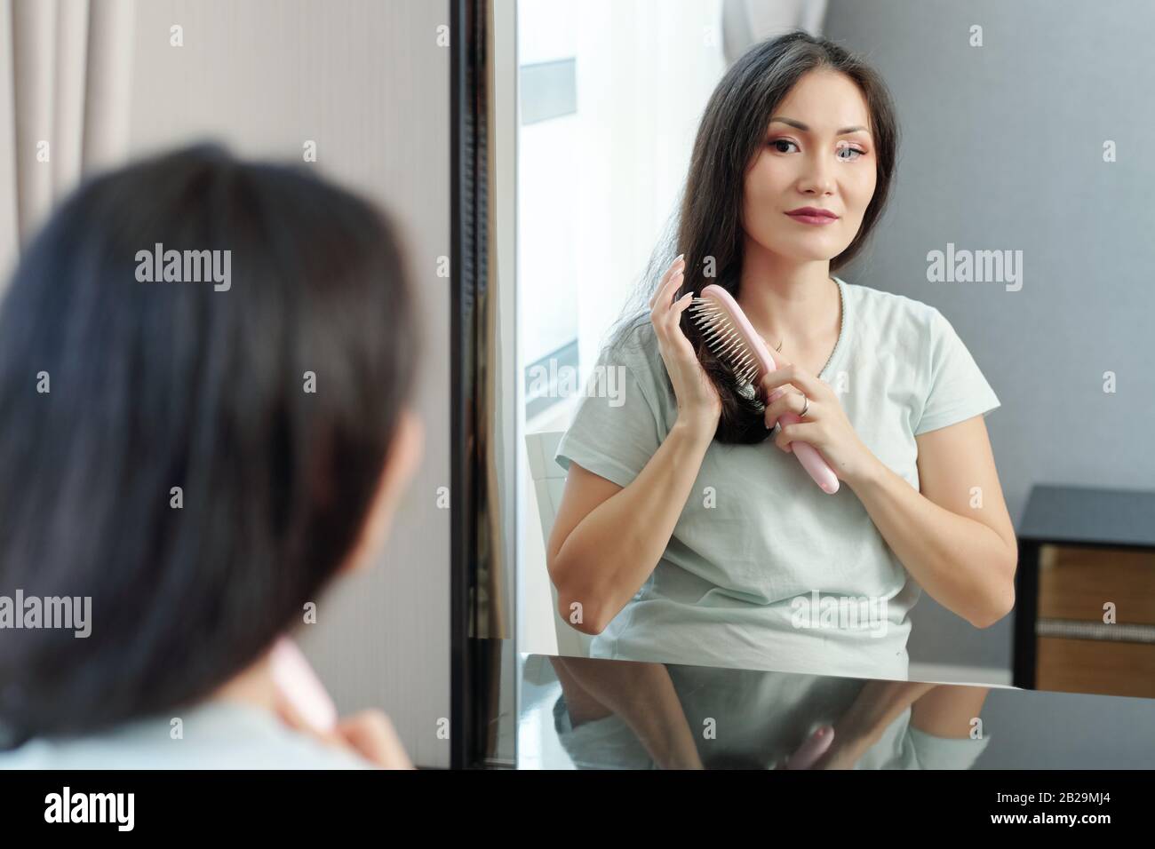 Beautiful young Vietnames woman sitting in front of mirror and enjoying brushing hair Stock Photo