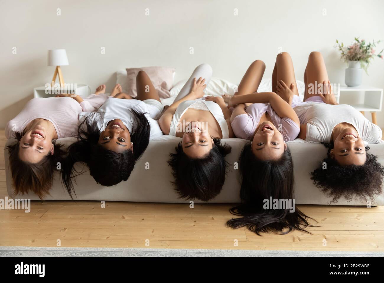 Happy diverse girls lying on bed upside down, having fun Stock Photo