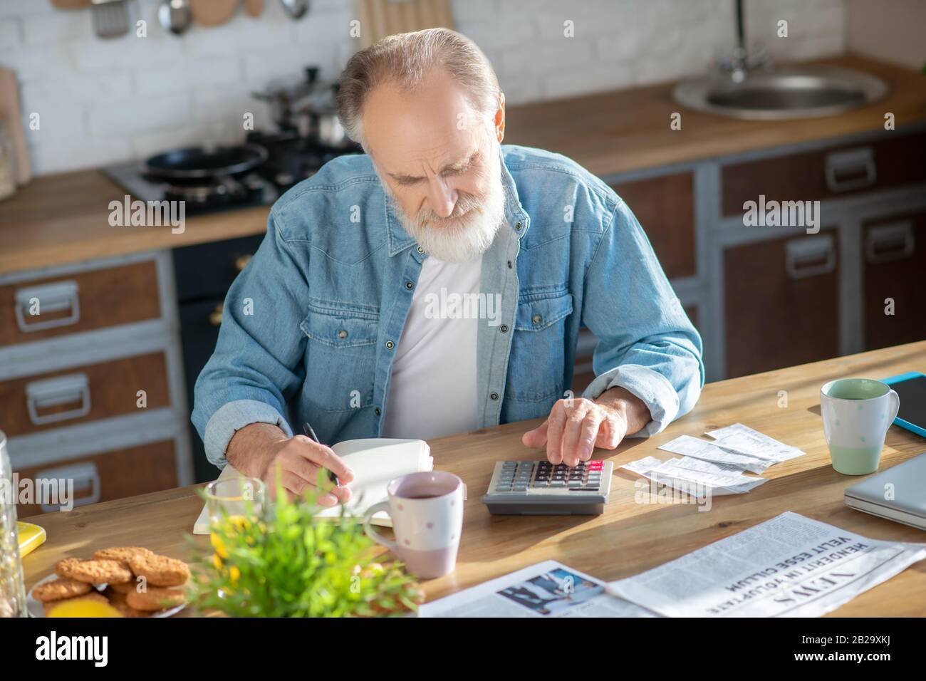 Bearded grey-haired man sitting at the table and making calculations Stock Photo
