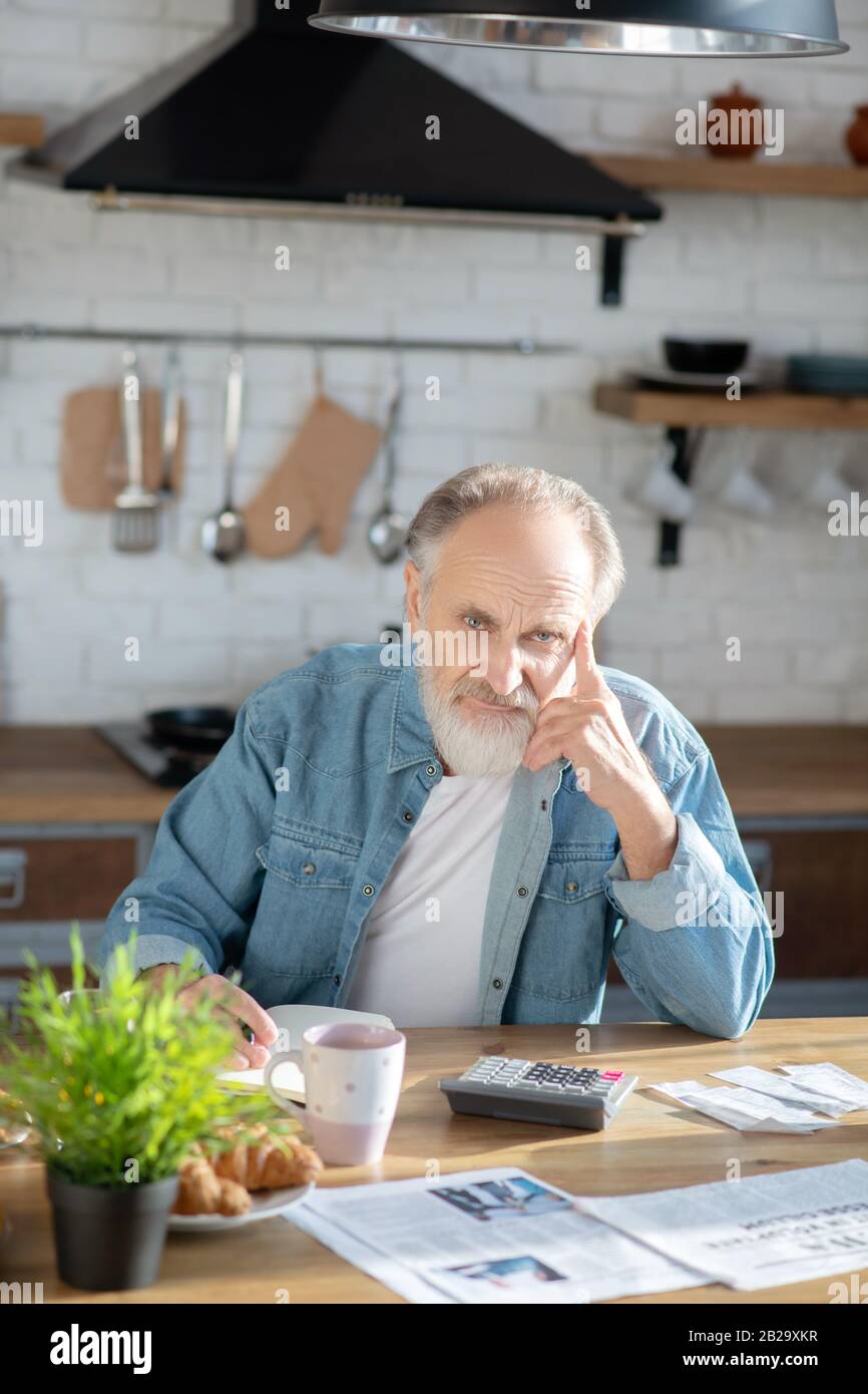 Bearded grey-haired man sitting at the table and looking thoughtful Stock Photo
