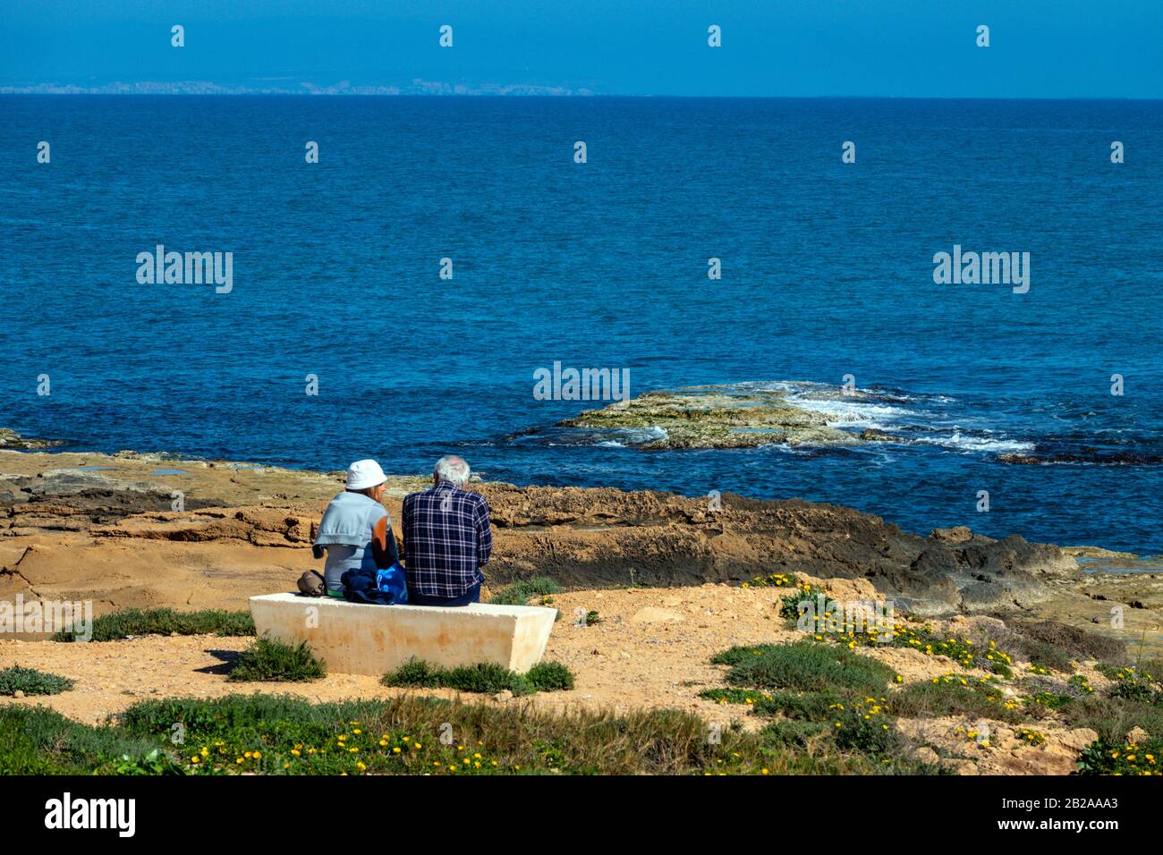 Two old people sitting on concrete bench looking at the ocean, Torrevieja, Costa Blanca, Spain Stock Photo