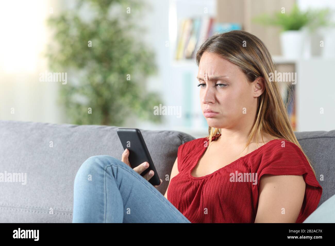 Confused woman after looking at her phone sitting in the couch at home Stock Photo