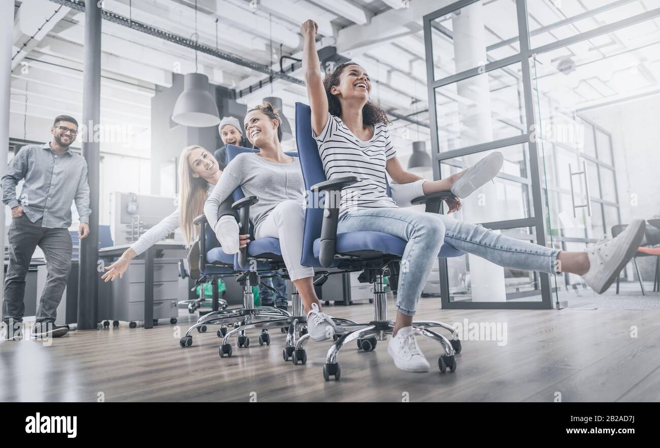 Young cheerful business people dressed in casual clothing are having fun on rowing chairs in a modern office. Happy team concept. Stock Photo