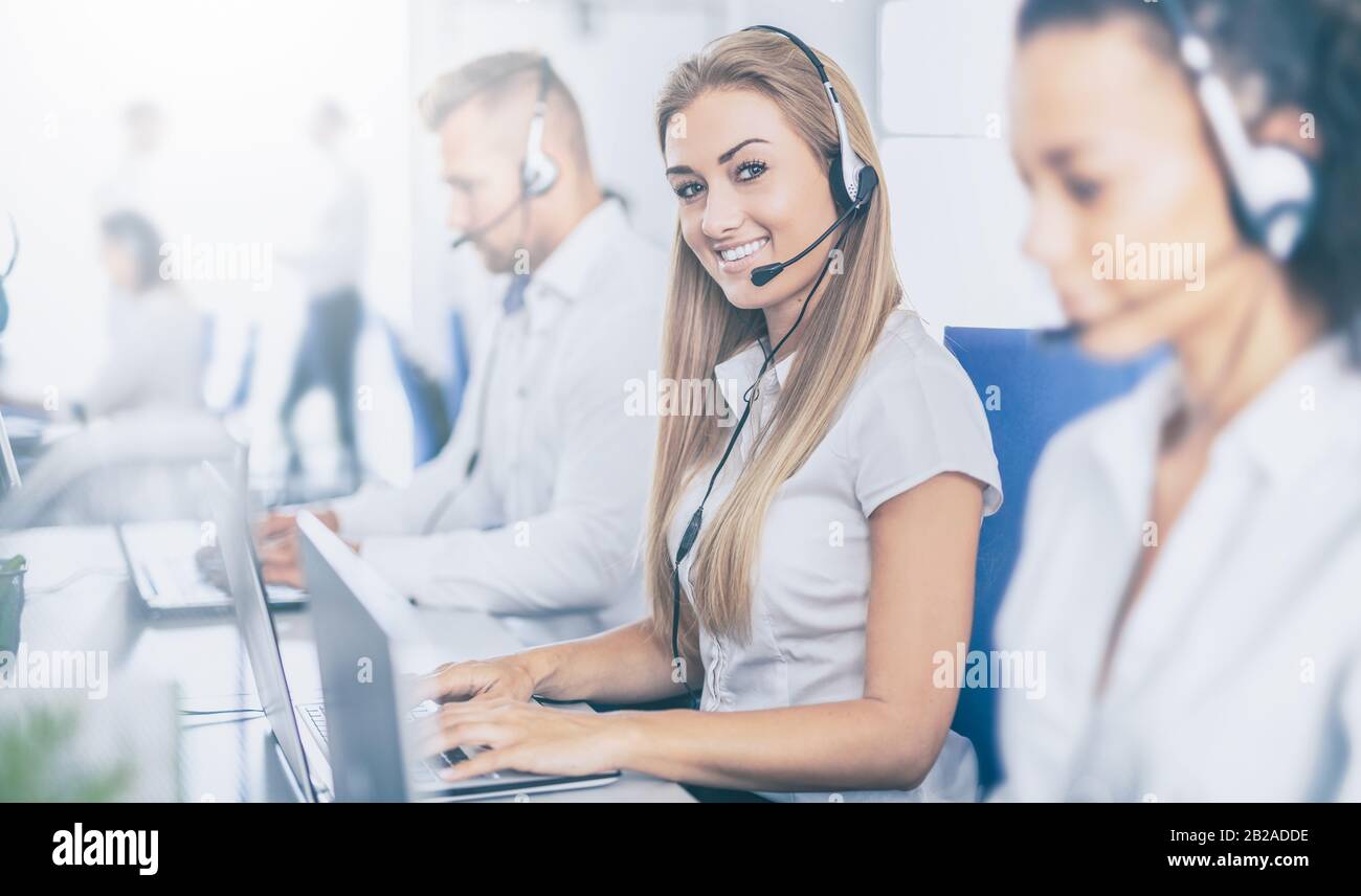 Call center worker accompanied by her team. Smiling customer support operator at work. Young employee working with a headset. Stock Photo