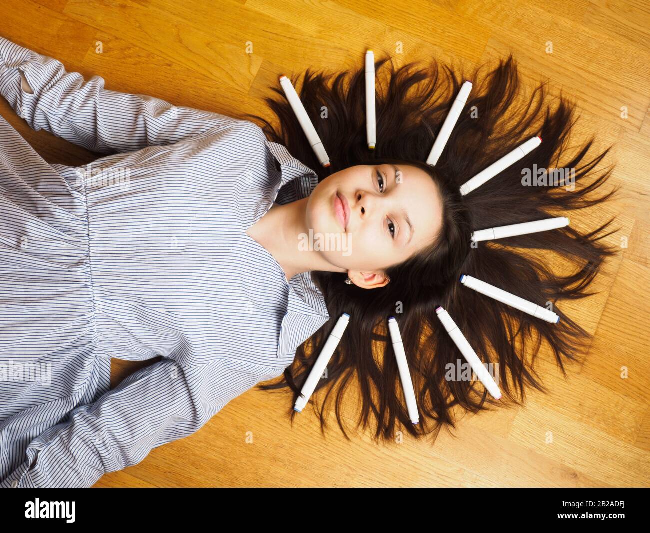 Young girl artist lies on a wooden floor in a light dress with her hair fluffy on which are felt-tip pens for drawing Stock Photo