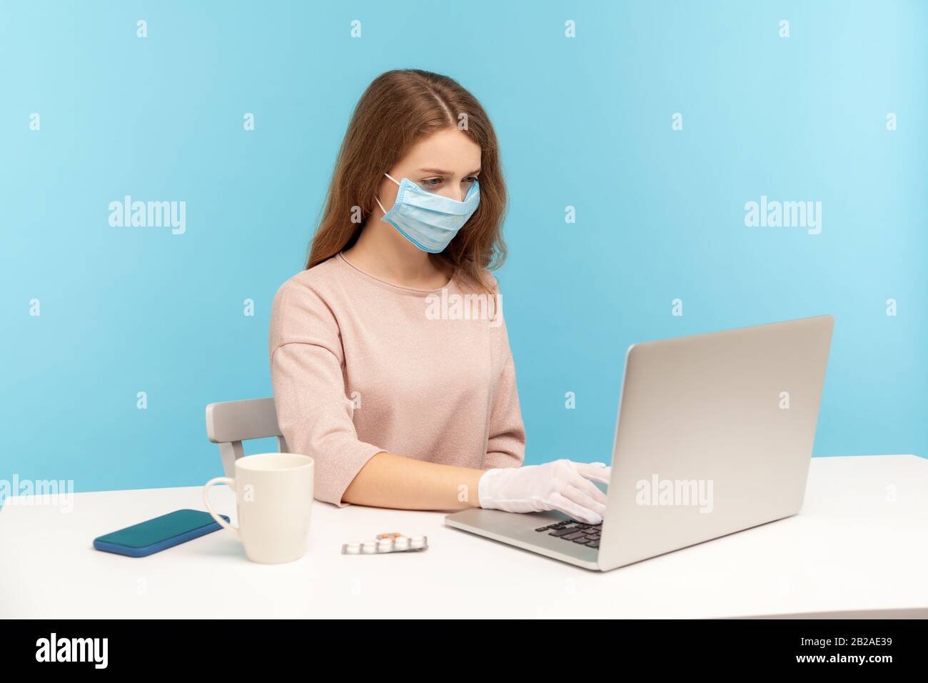 Office worker wearing facial mask and hygienic gloves while typing on laptop, protecting her hands and face to prevent contagious disease, viral infec Stock Photo