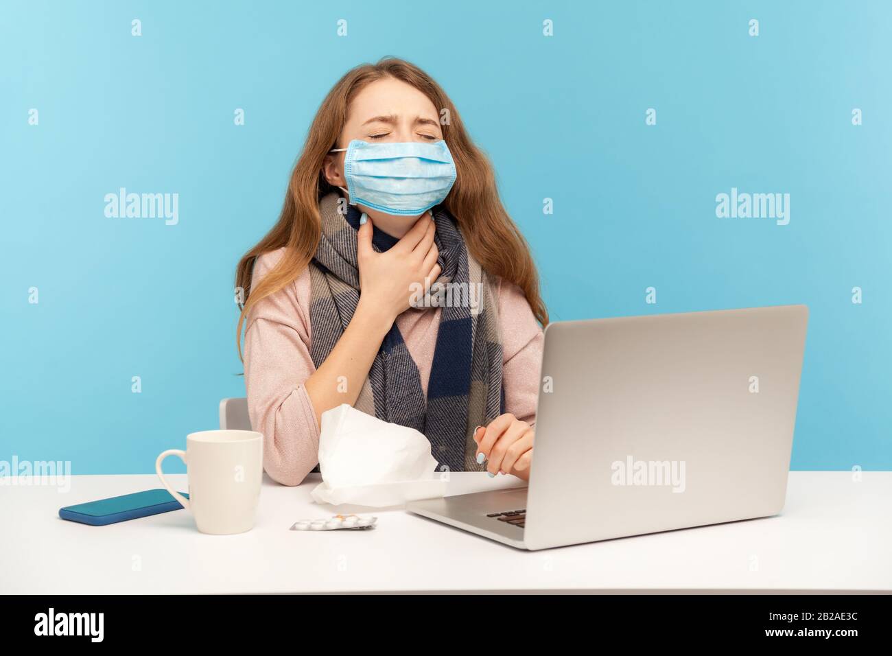 Sick girl in hygienic face mask sitting at desk with laptop, holding painful neck suffering sore throat, ill freelance woman with flu working from hom Stock Photo
