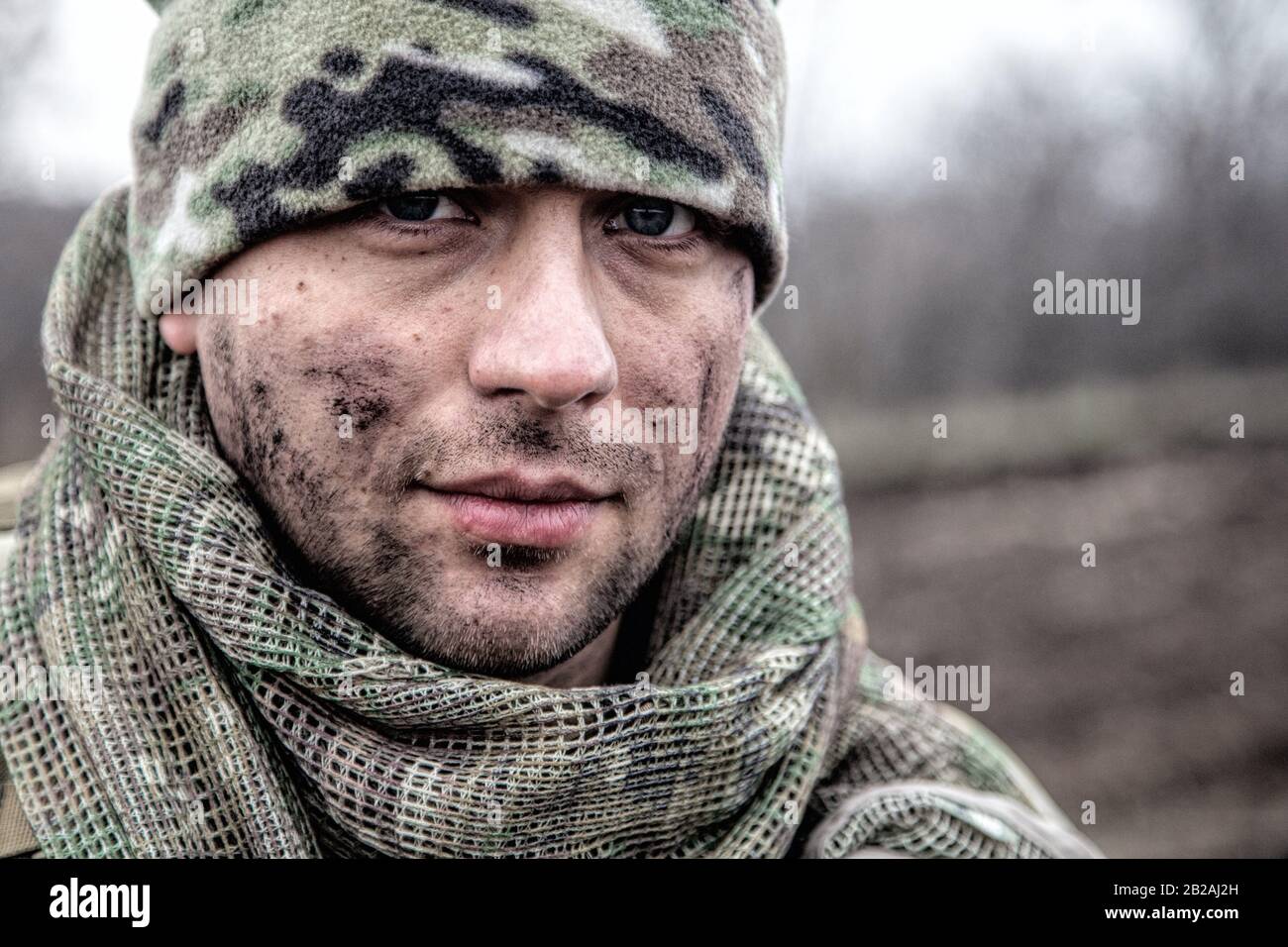 Portrait of soldier, modern combatant with dirty face, firearm replica, wearing camouflage uniform, beanie and masking cape on neck, standing on Stock Photo