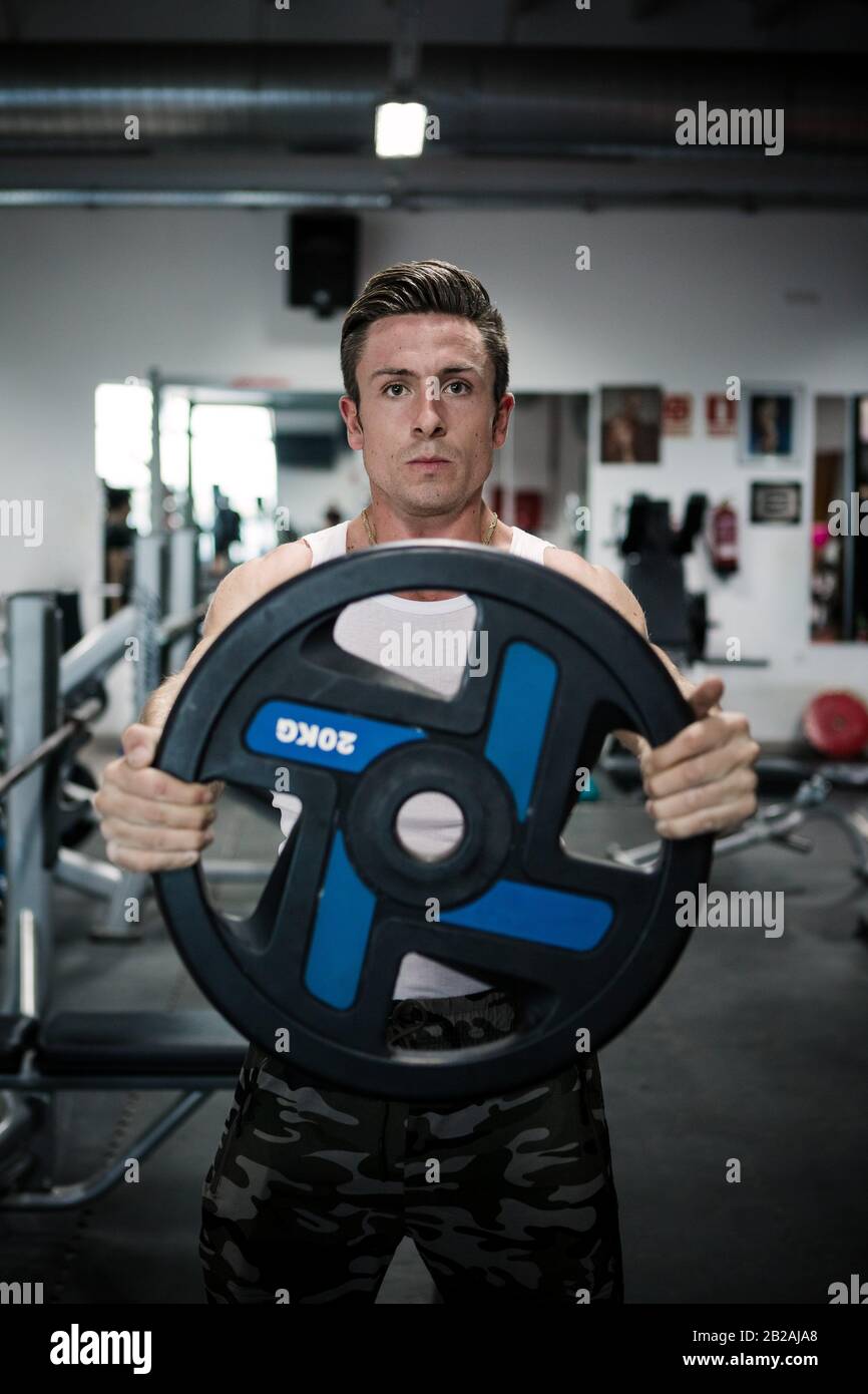 Determined muscular guy in sportswear doing exercise with heavy weight plate and looking at camera during workout in gym. Stock Photo