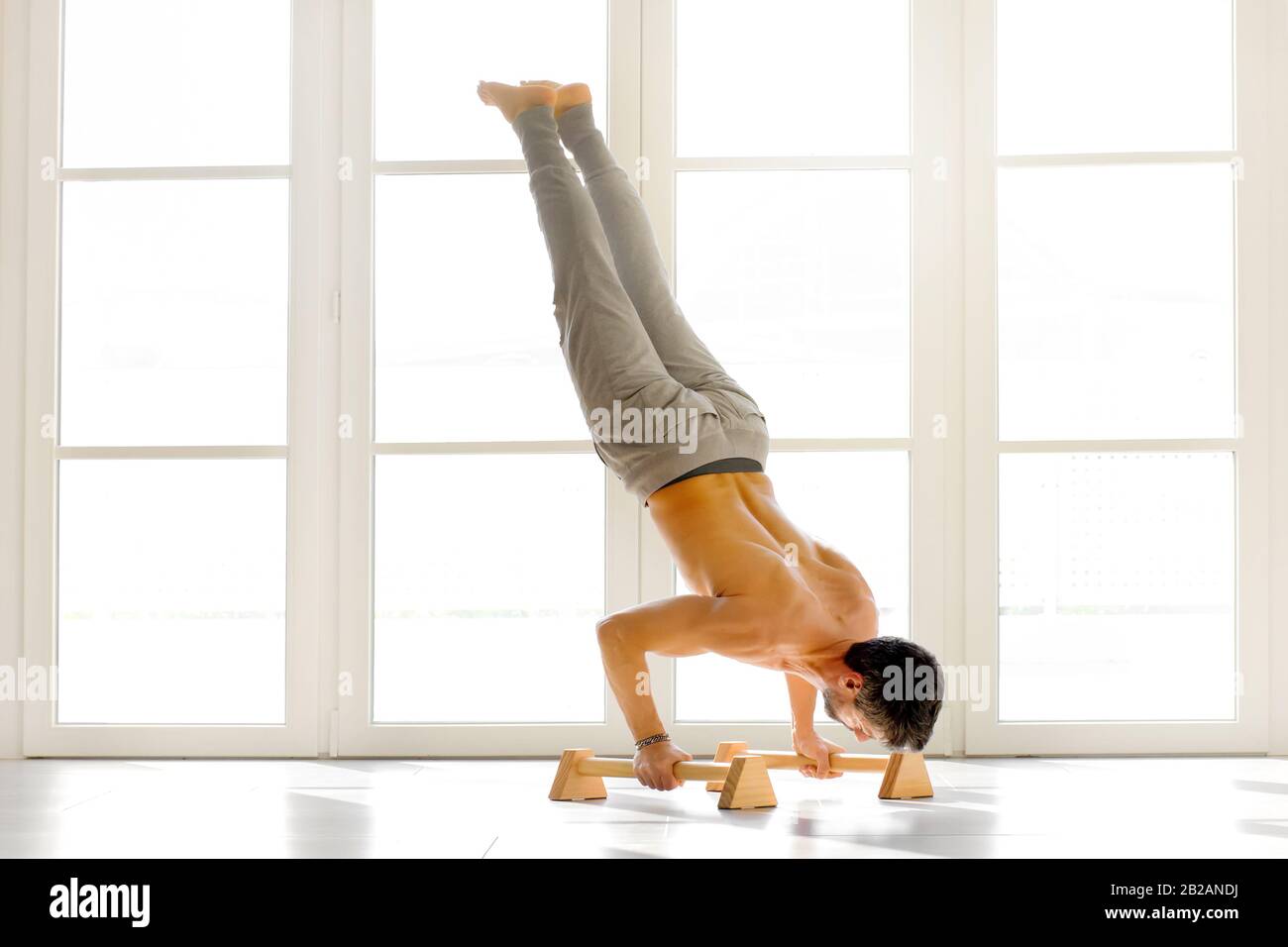 Muscular shirtless man doing power handstand push ups on low bars against bright white windows indoors Stock Photo