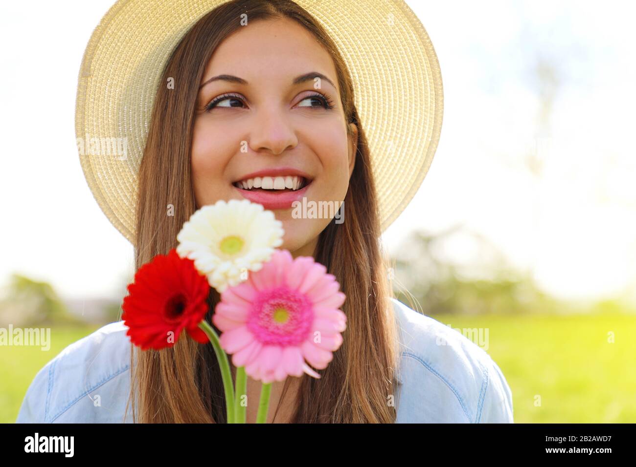 Spring girl holds flowers looking to the side outdoor. Beautiful smiling young woman with hat holding flowers in park and looks around. Stock Photo