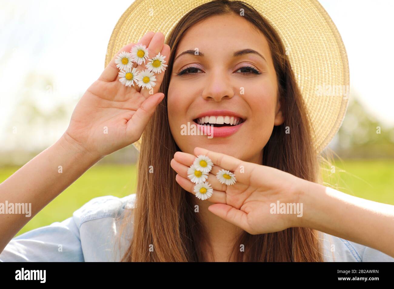 Close up portrait of a beautiful smiling girl with brown hair wearing hat and holds daisies between fingers looking at camera outdoors Stock Photo