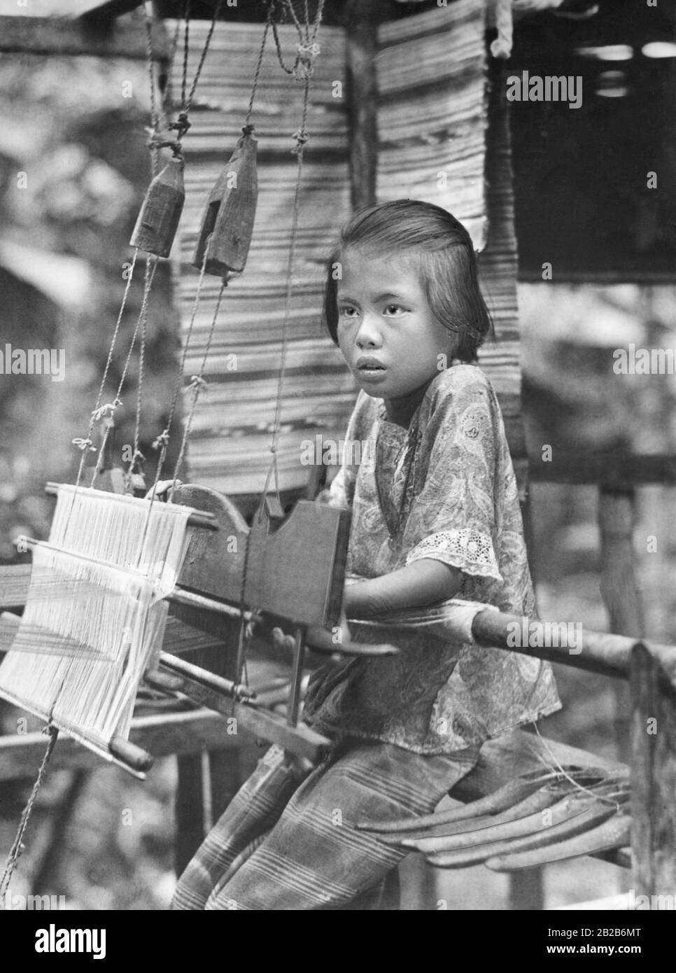 A little girl at the silk weaving mill in North Siam (today's Thailand). Laotian children learn to work at an early age. Silk weaving is an important home industry in this region. Stock Photo