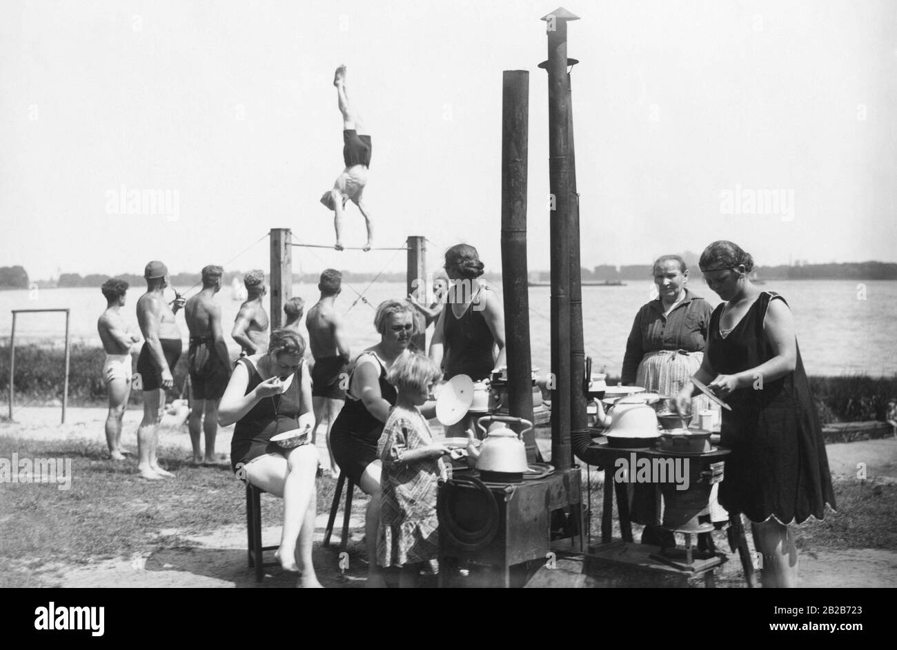 Campers gather around a cooking area with several ovens and prepare food in the Tegel tent camp at Lake Tegel. In the meantime, the men watch as one of them exercises on a horizontal bar. Stock Photo