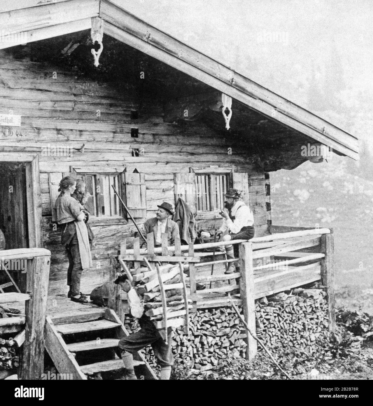 The picture shows a dairymaid with her child in her arms, greeting hikers who have settled on the forecourt of the hill farm. In the foreground a helper of the dairymaid is carrying firewood for the oven in the house on his back. The farm is located in the Krottenthal in the Mangfall Mountains in Upper Bavaria. Stock Photo