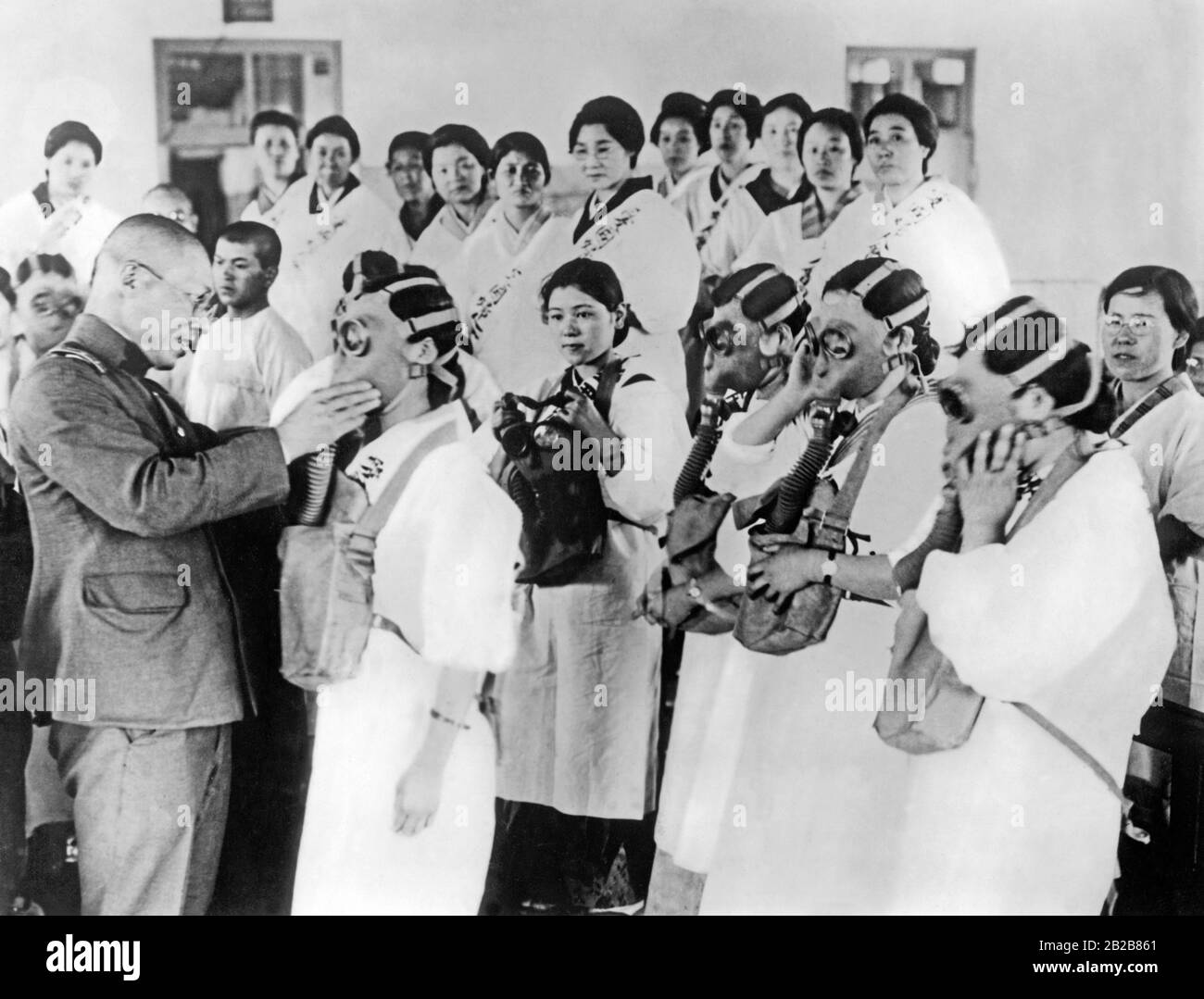 A Japanese soldier conducts a civil air-raid exercise with a group of women. He helps one of them to put on a gas mask. (undated picture, ca. 1930s) Stock Photo