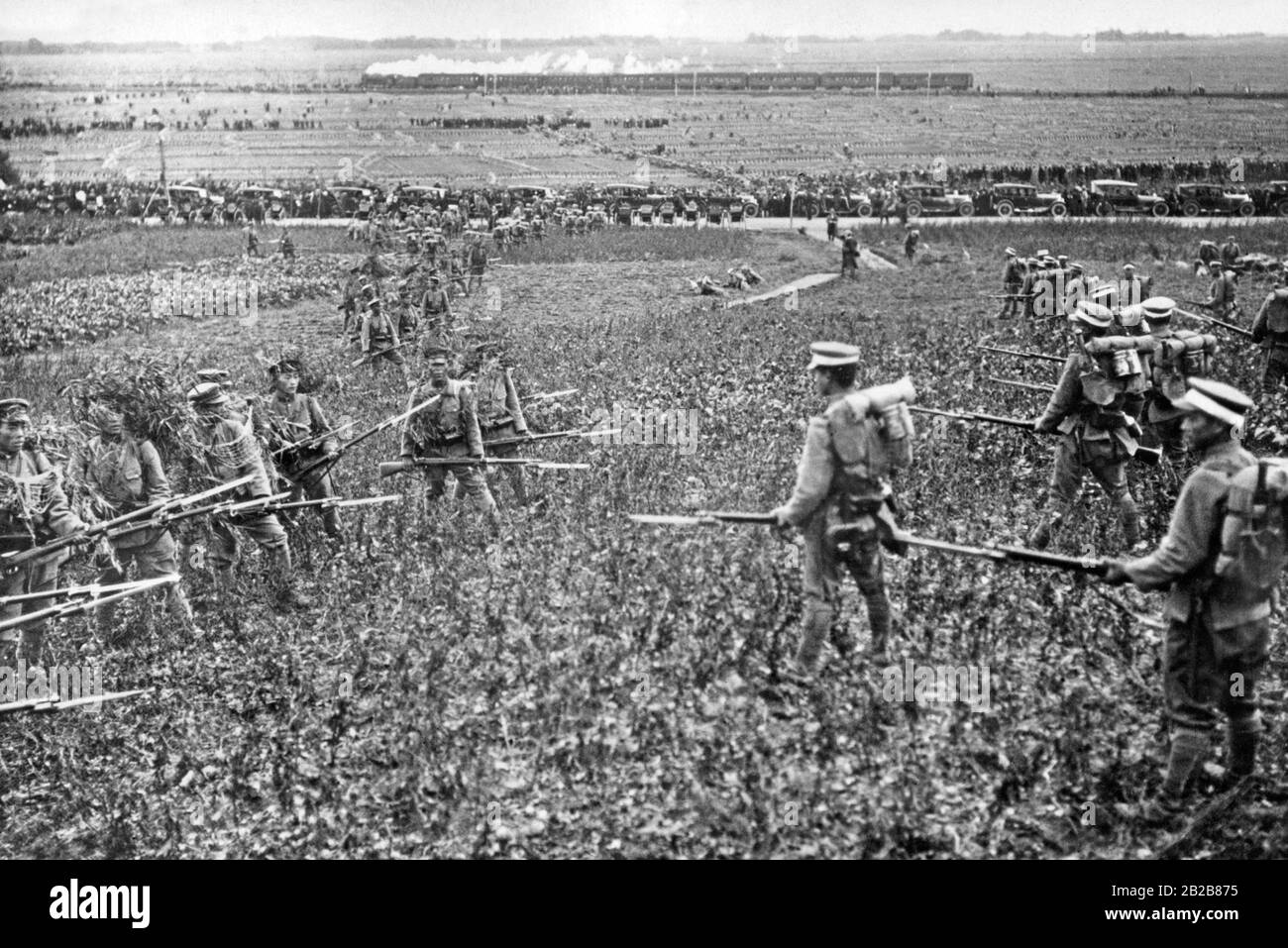 View over a wide field during an exercise maneuver of the Japanese military. In the foreground infantrymen of the enemy parties of the exercise are facing each other. They carry rifles with bayonets attached. Behind them is a country road lined with carts and cars. In the background a train is running. (Undated photo, ca. 1930s) Stock Photo