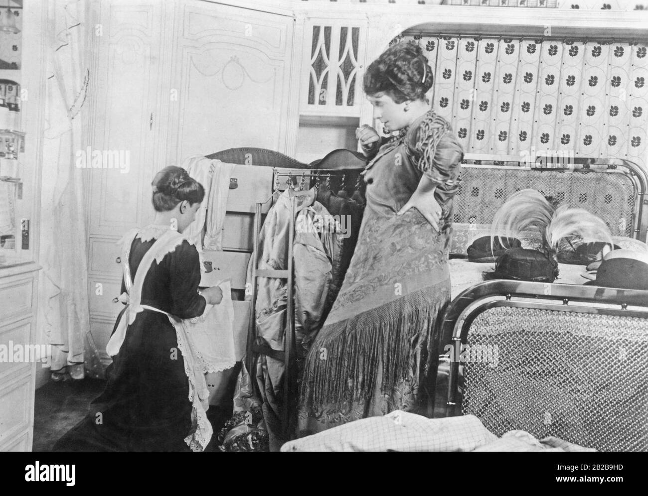 Elegant lady gives orders to her maid as she packs her suitcase. Behind