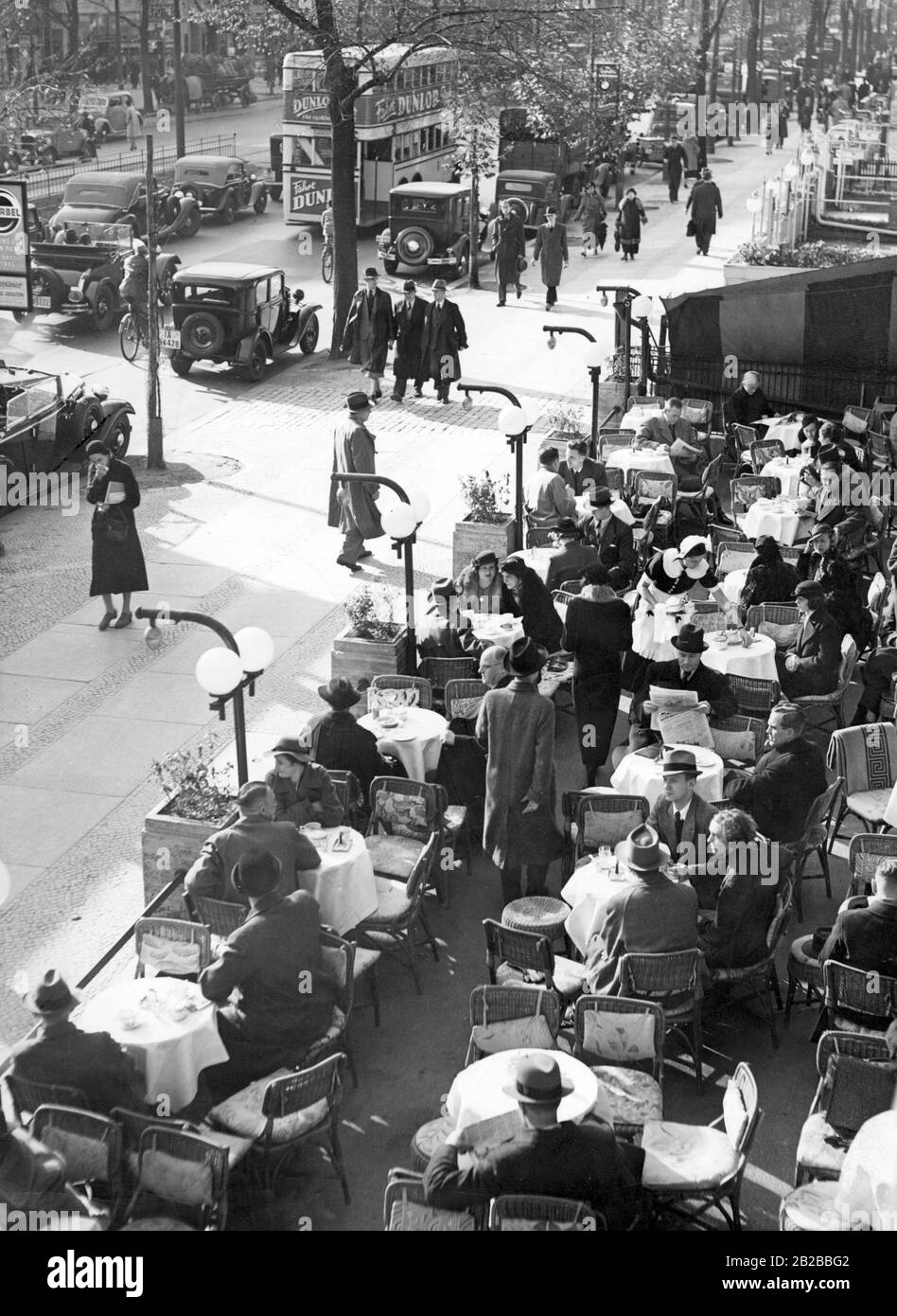 People sit on the terrace of Cafe Kranzler on the Kurfuerstendamm in Berlin. The outside area of the cafe is bordered by flower troughs, which are illuminated. On the left there is a double-decker bus, typical for Berlin. Stock Photo