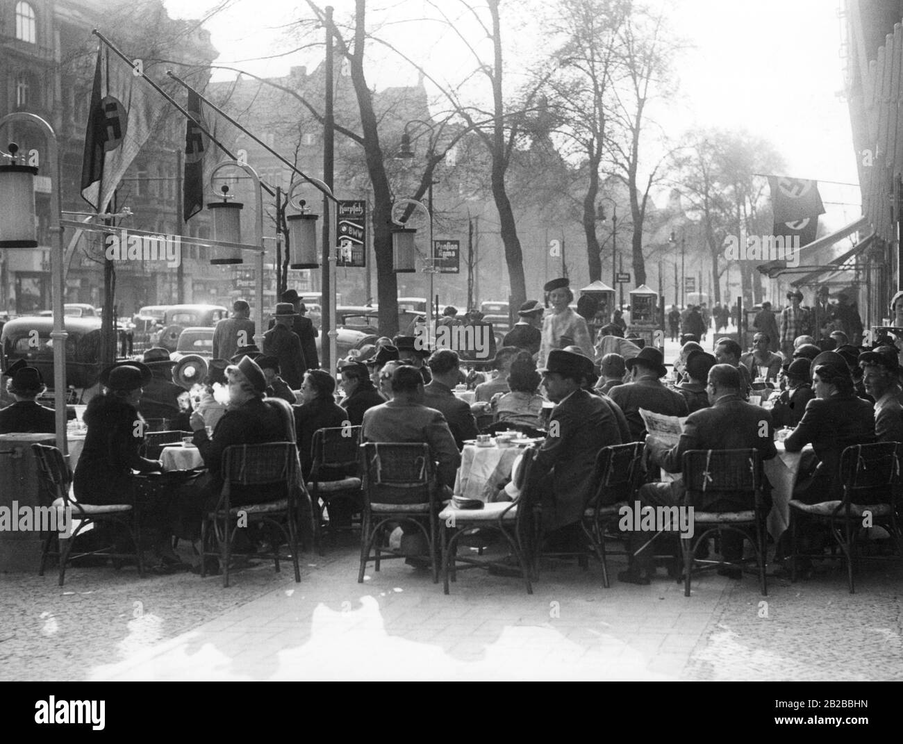People sit in a cafe on the sidewalk at Kurfuerstendamm in Berlin. Swastika flags are hoisted on the right on the house and on the left on the street lamps. Stock Photo