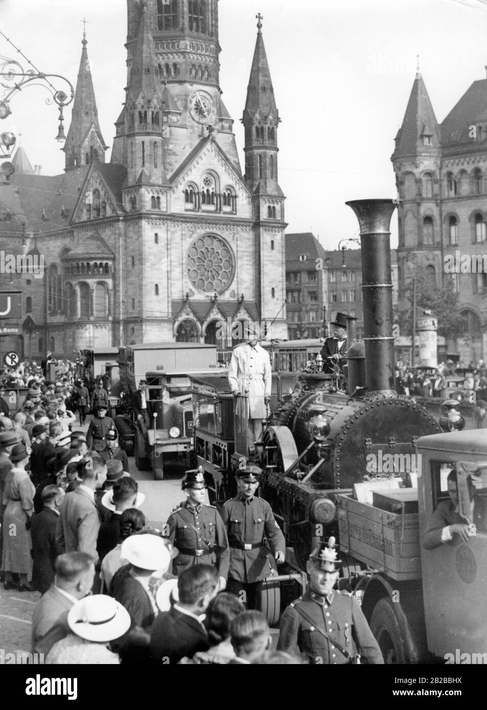 The replica of the first German locomotive 'Adler' is transported to the exhibition halls at Kaiserdamm in Berlin. The truck with the locomotive passes in front of the Kaiser Wilhelm Memorial Church, which can be seen in the background. Many people stand on the roadside. An SA man in white coat is standing on the locomotive. The train is accompanied by SS men with swastikas and policemen. Stock Photo