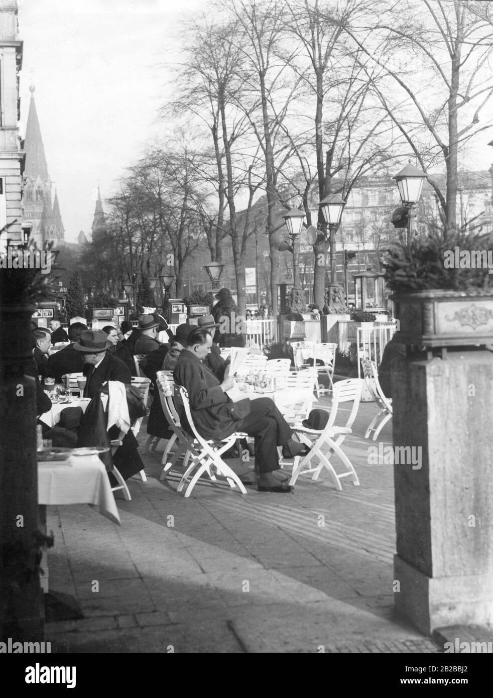 Guests sit in a street cafe in Berlin on the Kurfuerstendamm. It is photographed through two columns with plant troughs on them. He wears gaiters over his shoes.  In the background the Kaiser Wilhelm Memorial Church can be seen. Stock Photo