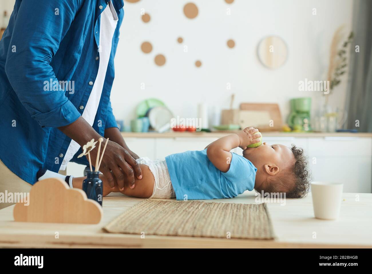 Side view at unrecognizable father changing diaper to cute African-American toddler eating apple, copy space Stock Photo
