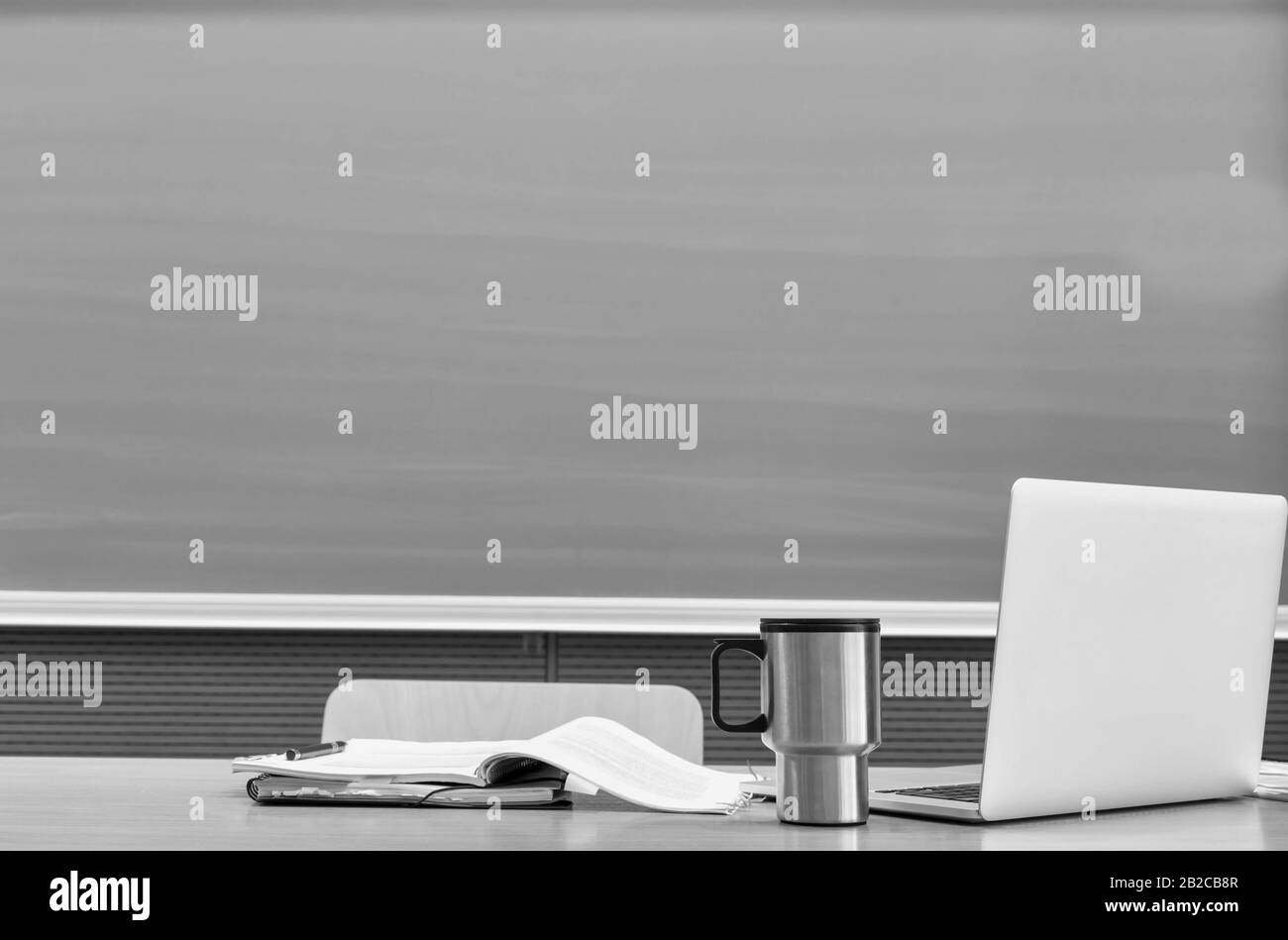 Black and white photo of laptop on professor desk with tumbler and book against black board in classroom Stock Photo