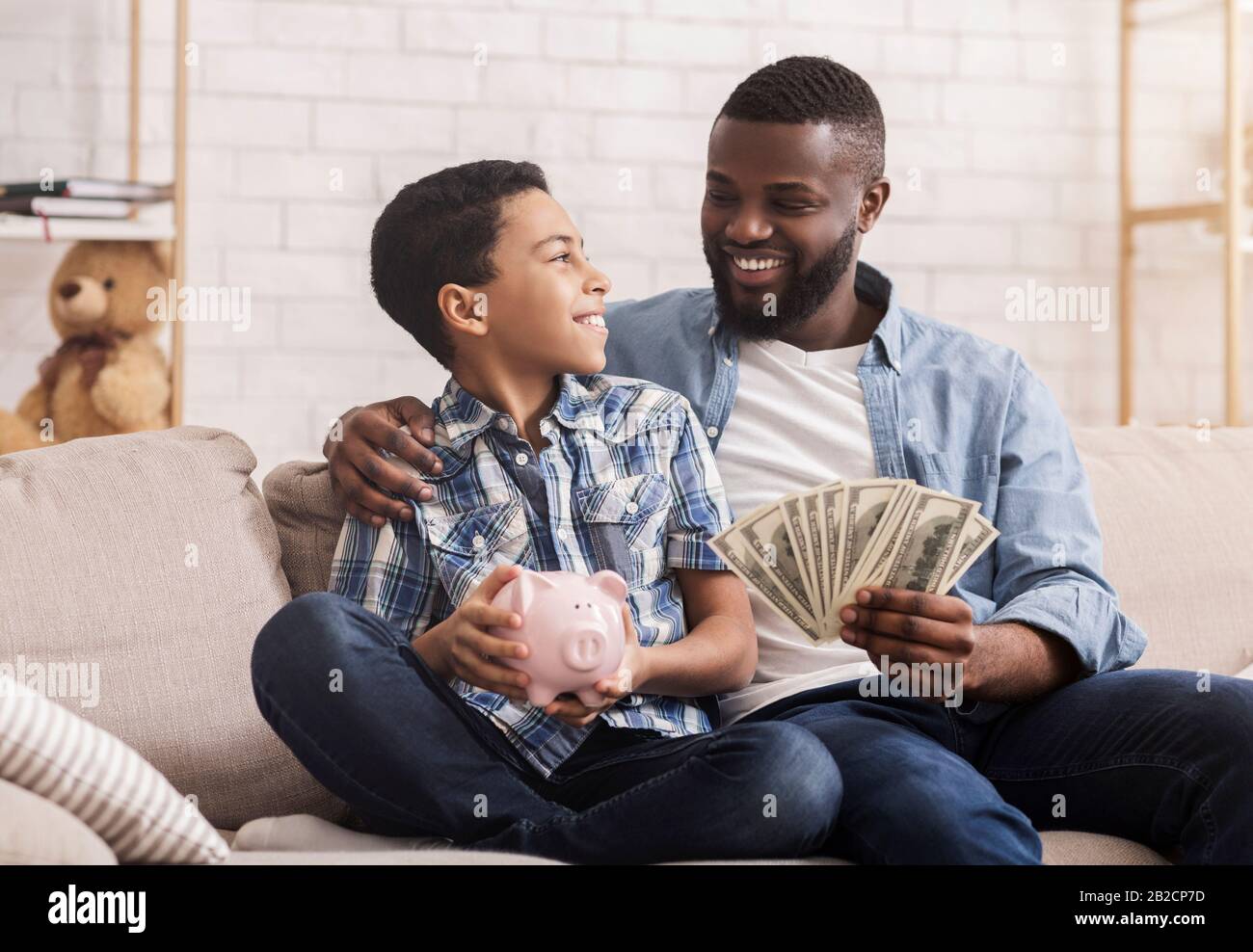 Father And Son Sitting On Couch With Cash And Piggy Bank Stock Photo