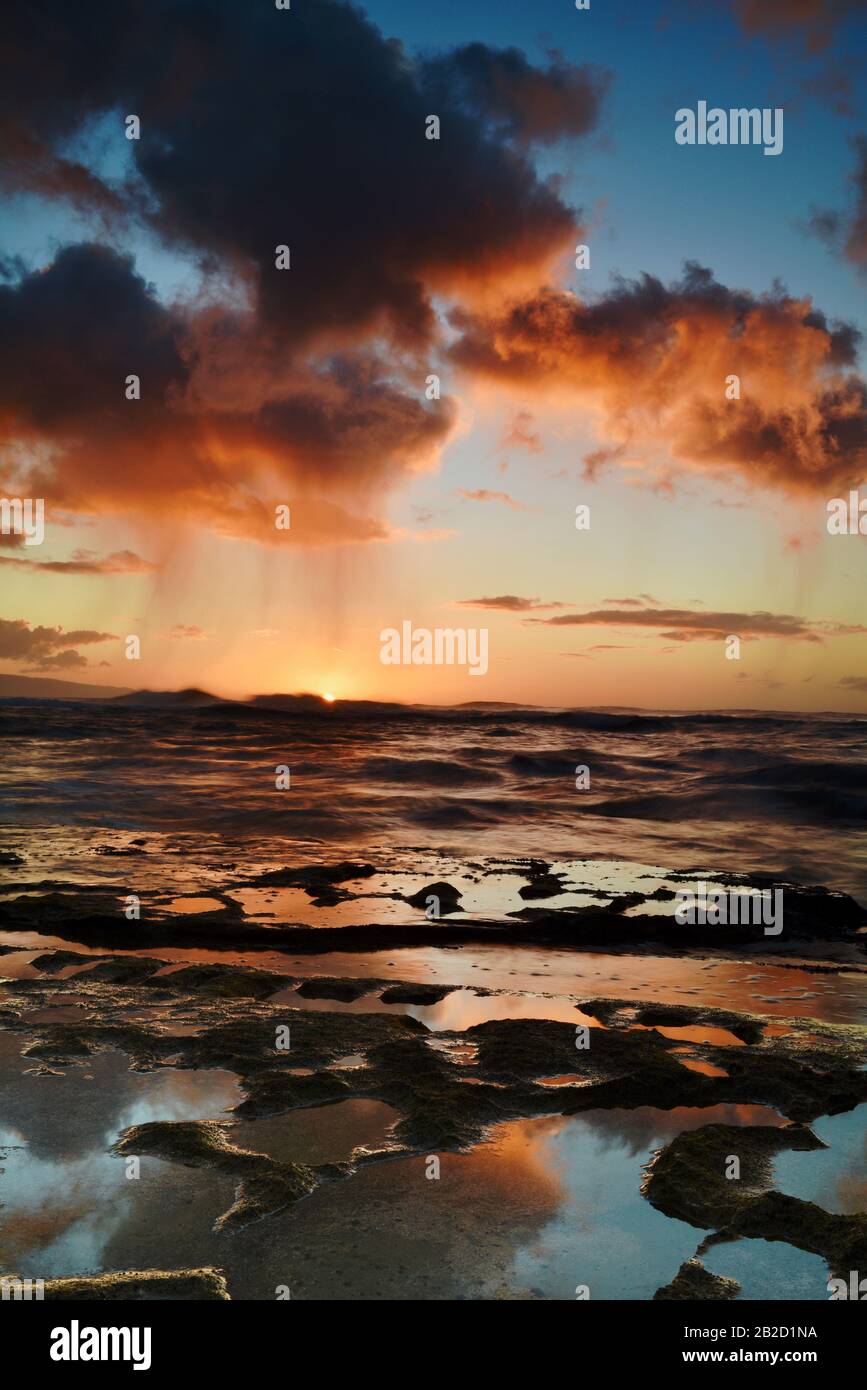 Dramatic sunset with rain showers falling from storm clouds over ocean, reflection in volcanic rocks, on the North Shore of Oahu, Haleiwa, Hawaii, USA Stock Photo