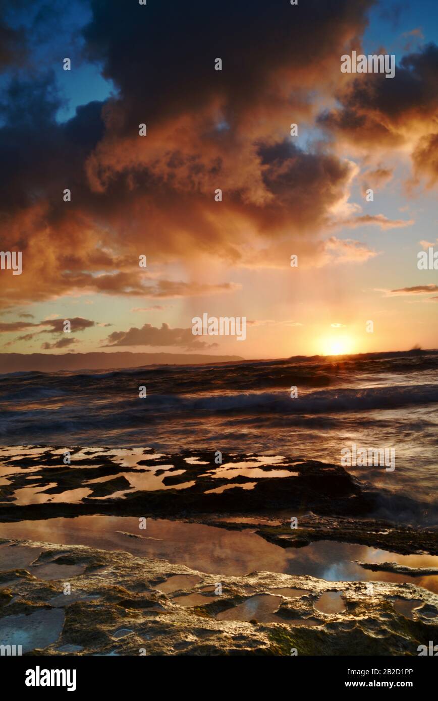 Dramatic sunset with rain showers falling from storm clouds over ocean, reflection in volcanic rocks, on the North Shore of Oahu, Haleiwa, Hawaii, USA Stock Photo