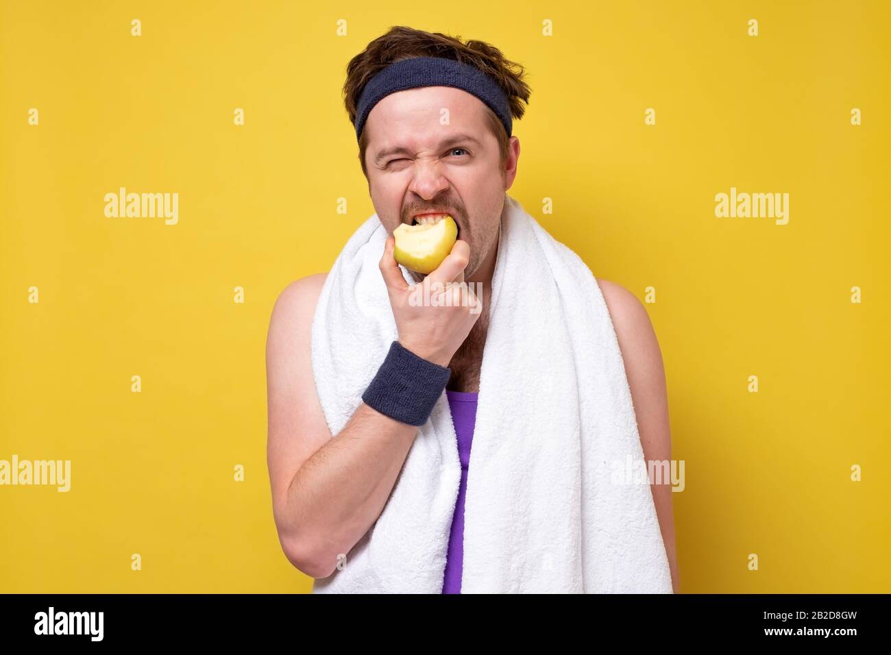 Fitness man with towel eating apple isolated on a yellow background. Healthy diet for sportsman. Stock Photo