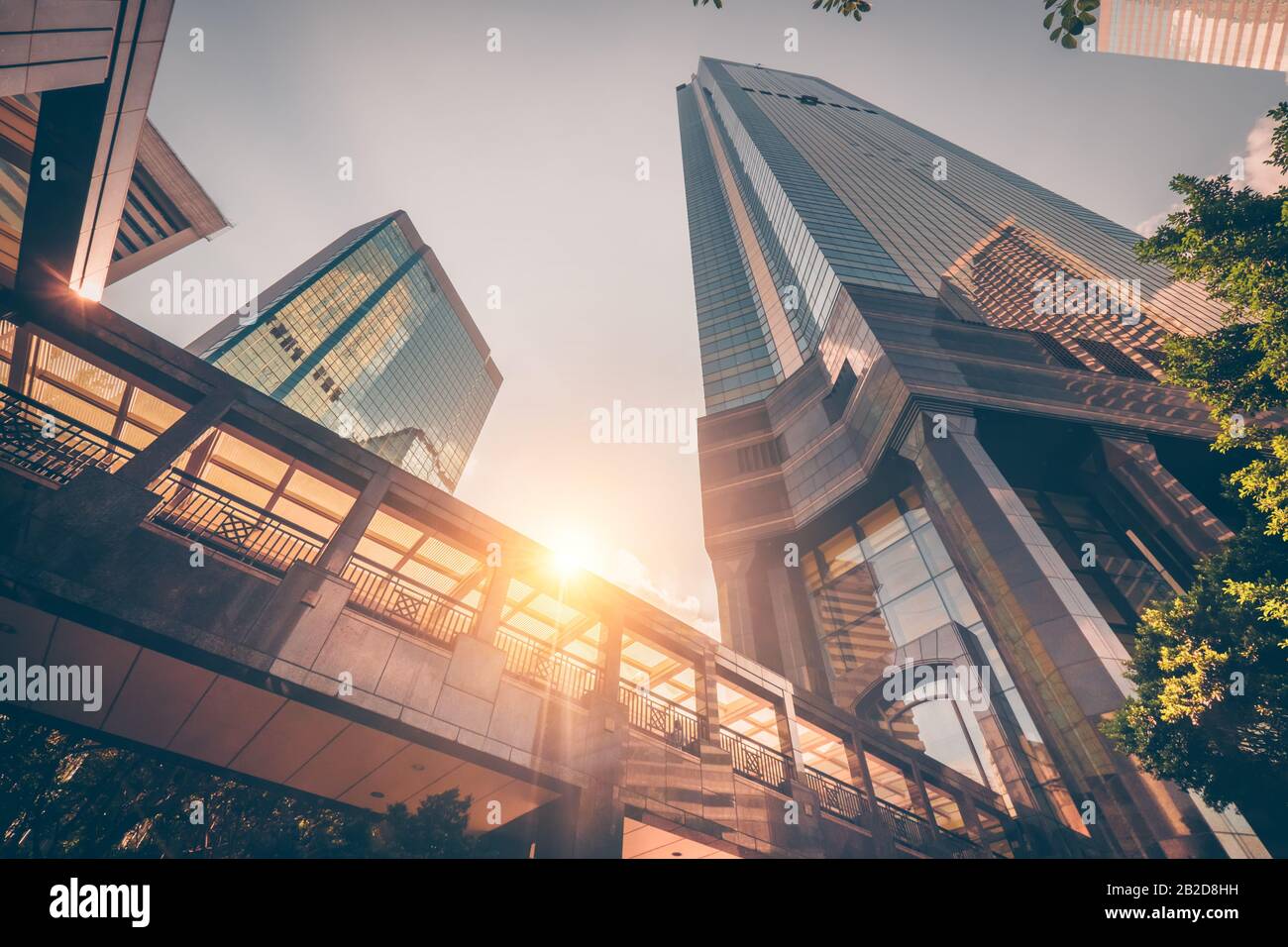 Abstract futuristic cityscape view with modern skyscrapers. Sun shines in the sunset sky, reflecting in glass of the footbridge. Urban architecture ba Stock Photo