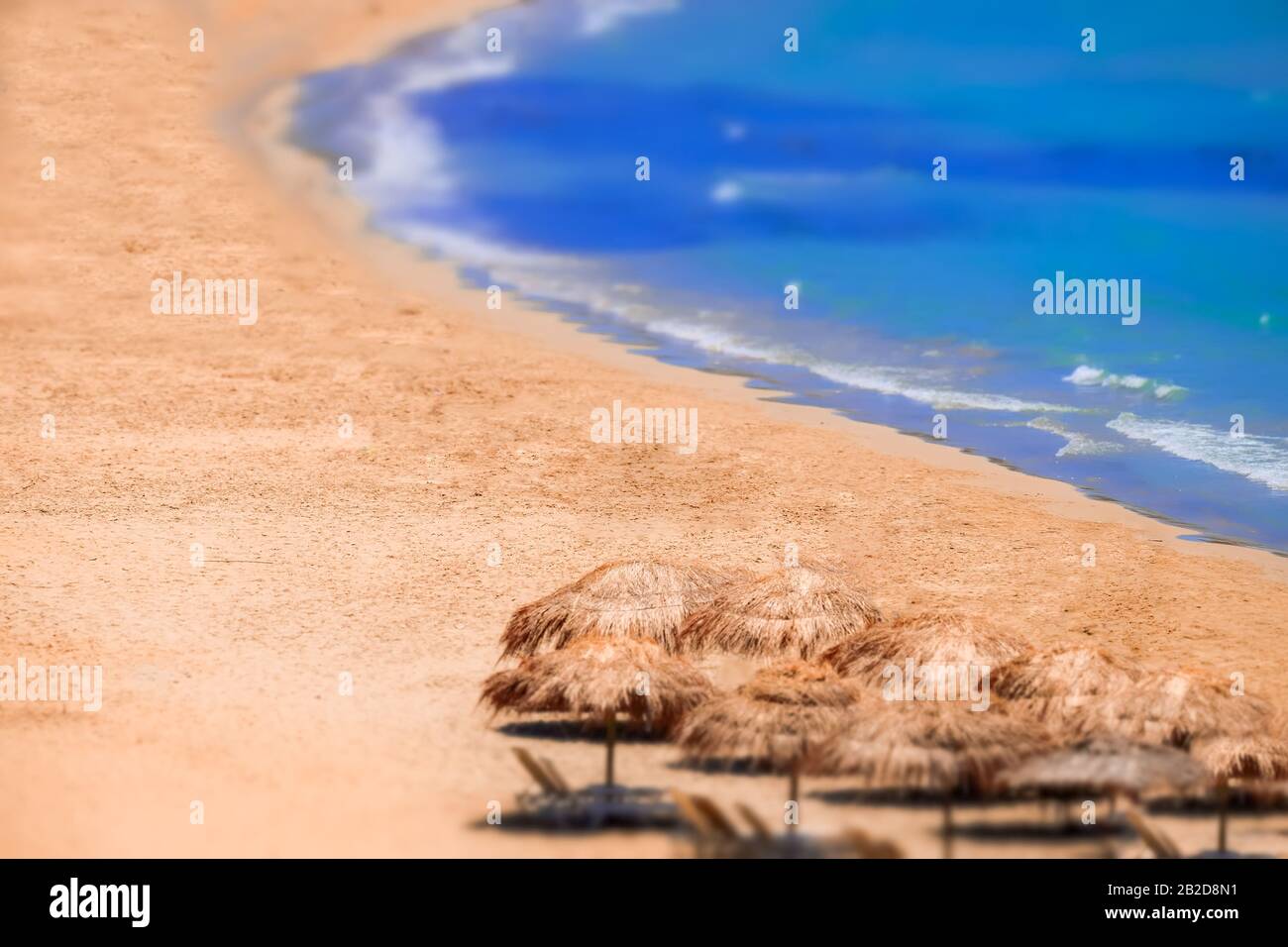 Tilt shift lens blur. Abstract view of amazing beach landscape with straw umbrella near azure sea. Mediterranean paradise in Greece. Travel background Stock Photo