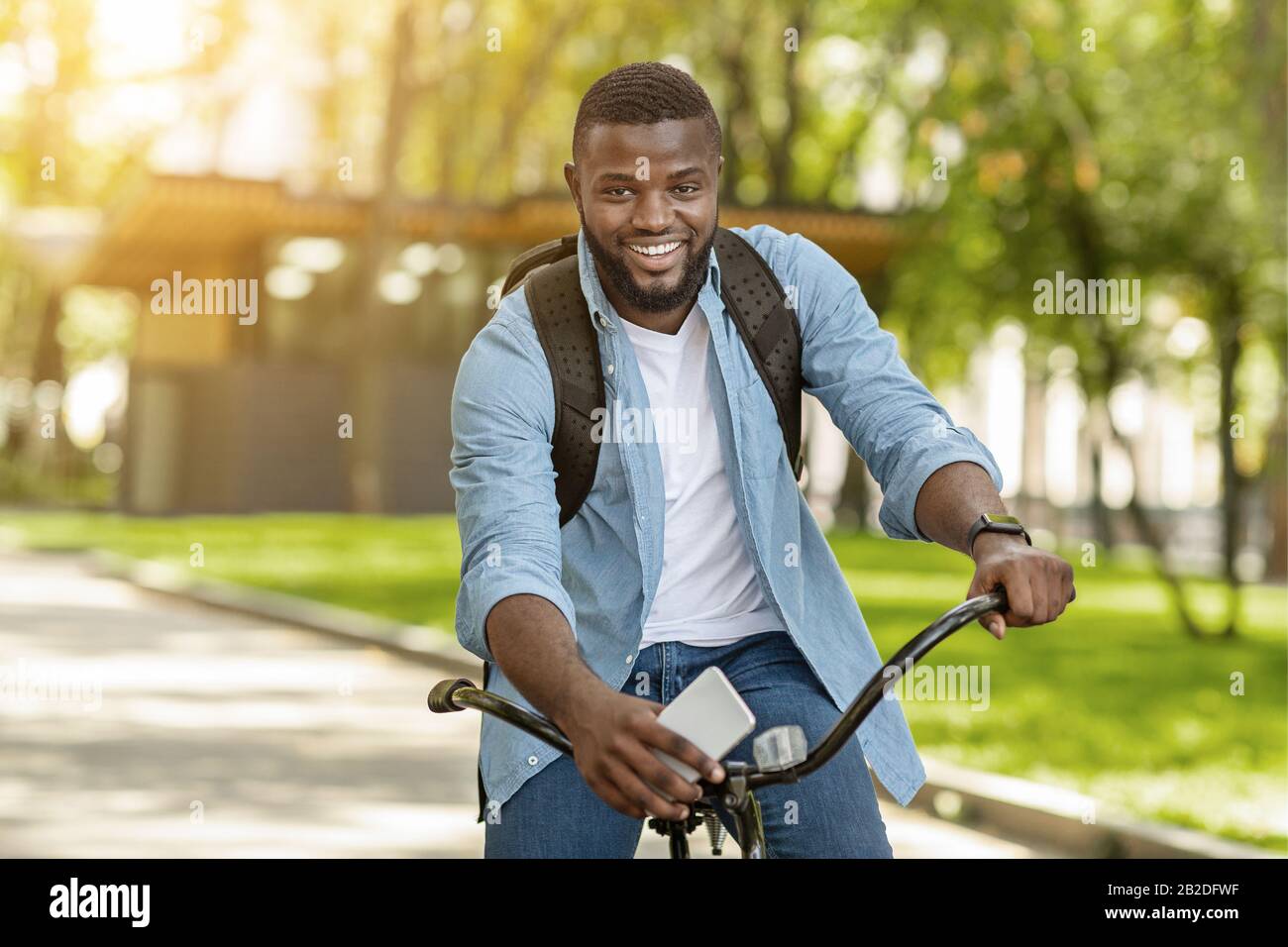Handsome Black Man With Backpack And Smartphone Riding Bicycle, Posing Outdoors Stock Photo