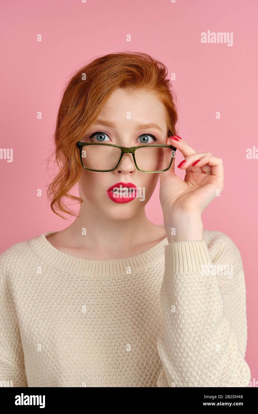 A red-haired girl in a white sweater with red lipstick stands on a pink background, adjusting glasses and looking at the camera Stock Photo