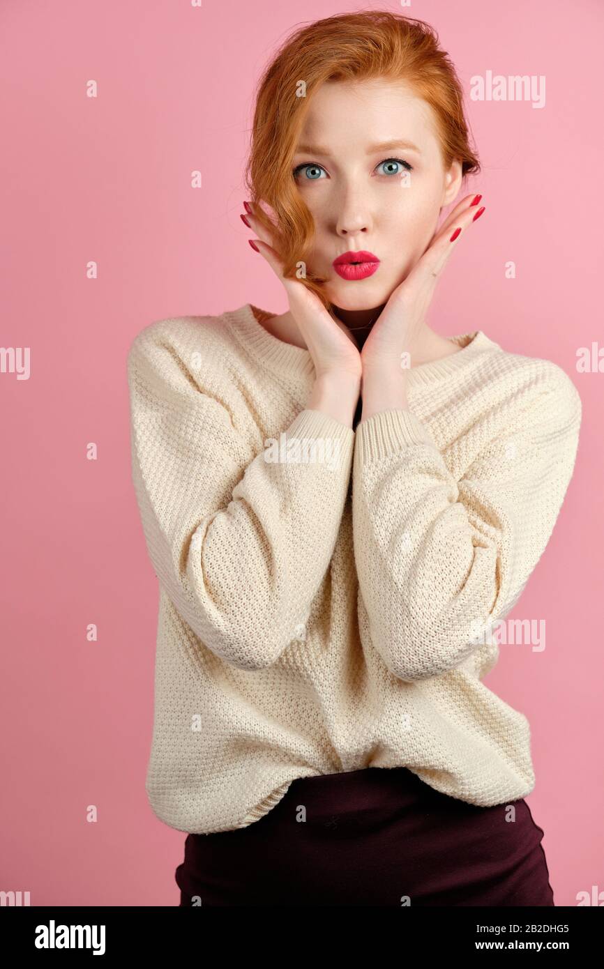 A red-haired girl in a white sweater with red lipstick stands on a pink background holding her hands to her face. Stock Photo