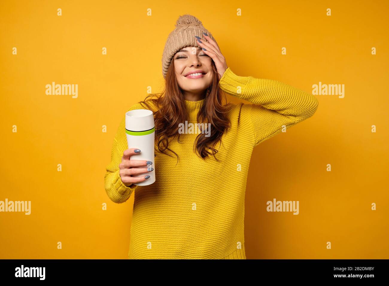 A brunette in a yellow sweater and hat is standing on a yellow background, smiling pretty with a thermocup in her hands. Stock Photo