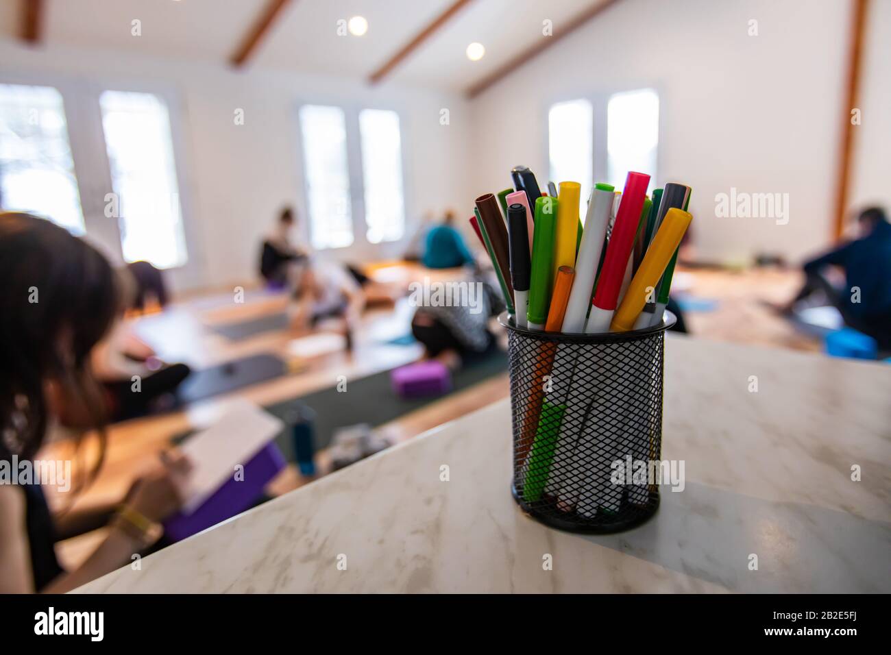 The small basket with multi-colored pencils. A little group of people taking a part in the yoga class at the background. The multi-colored handles Stock Photo