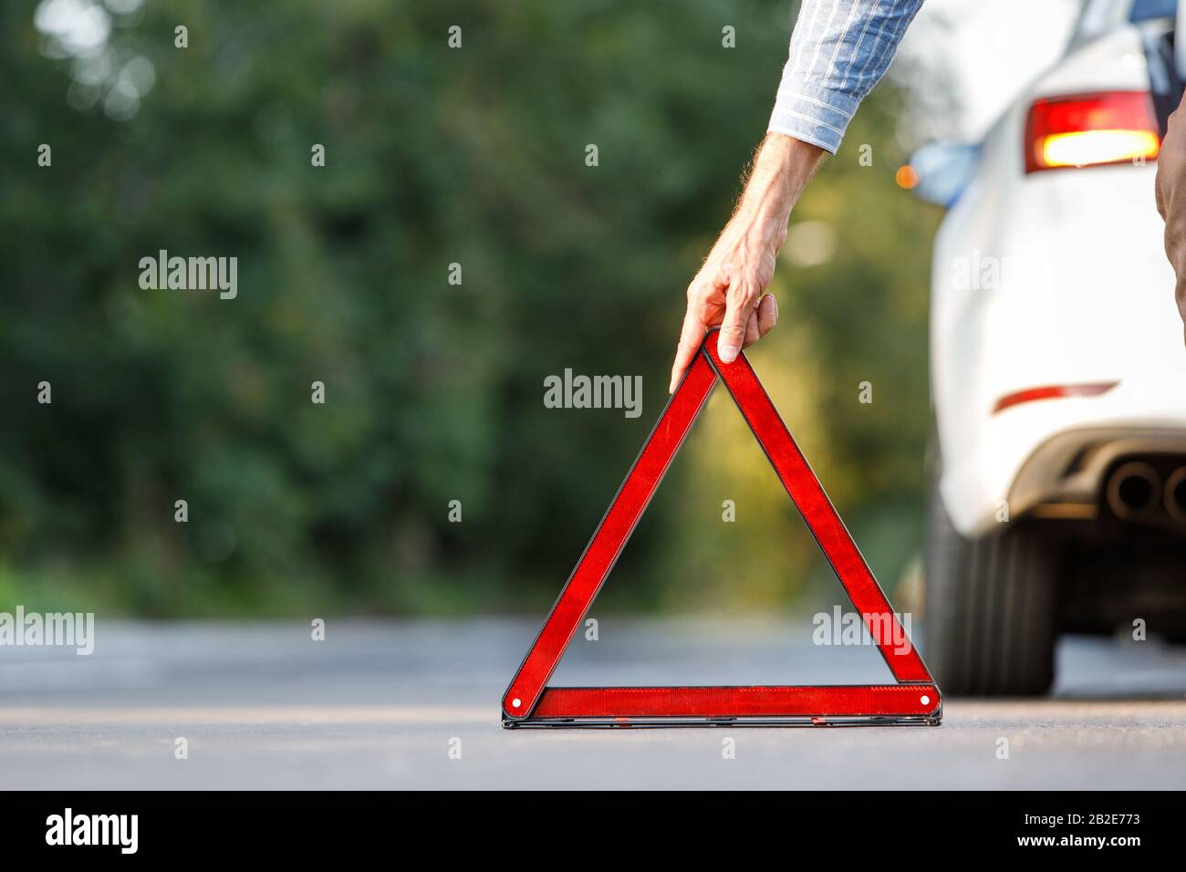 Close up of man driver putting red warning triangle/emergency stop sign behind his broken car on the side of the road, copy space and blurred backgrou Stock Photo